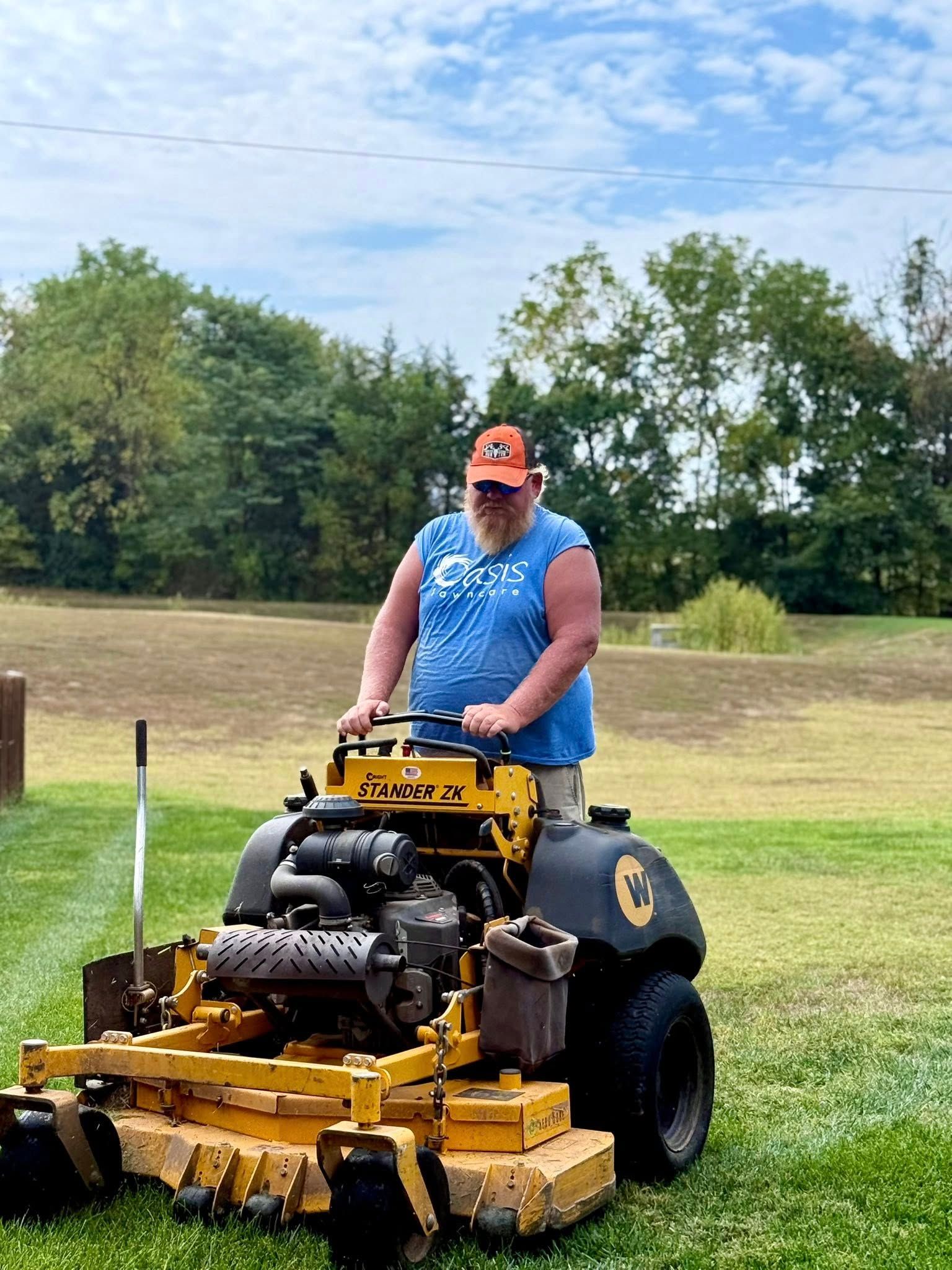 Man on a yellow riding lawnmower mowing grass in a grassy field with trees in the background.