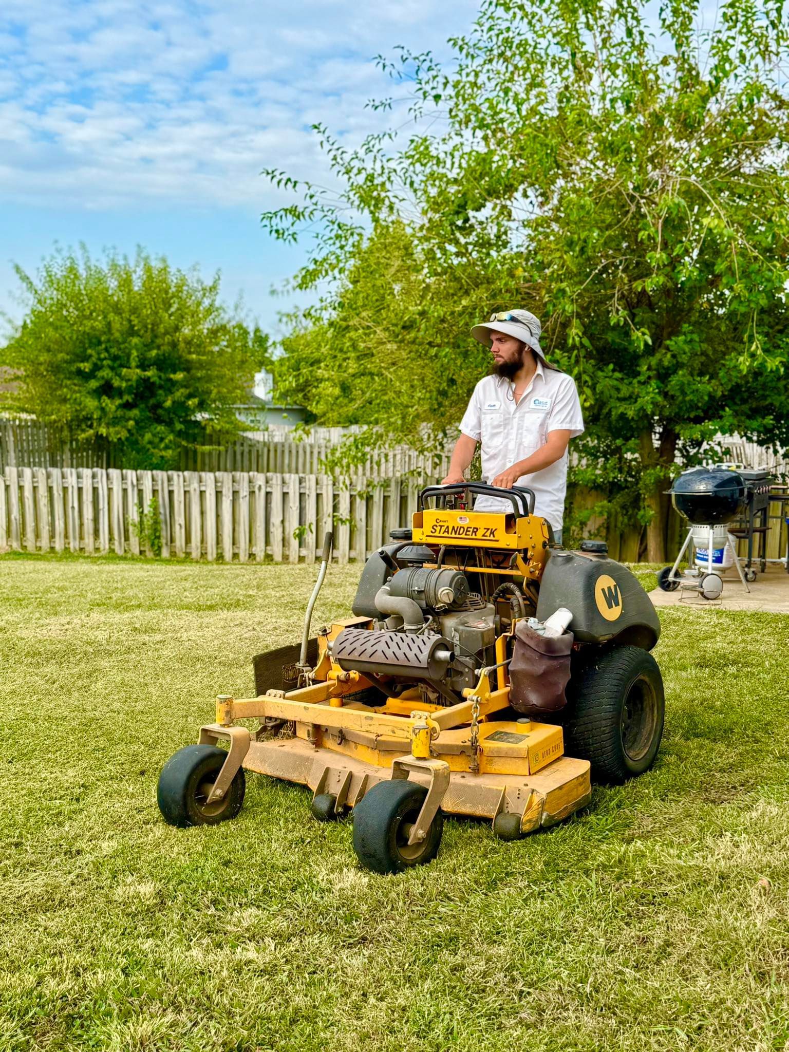 Person on yellow lawnmower in a yard with trees and a fence.