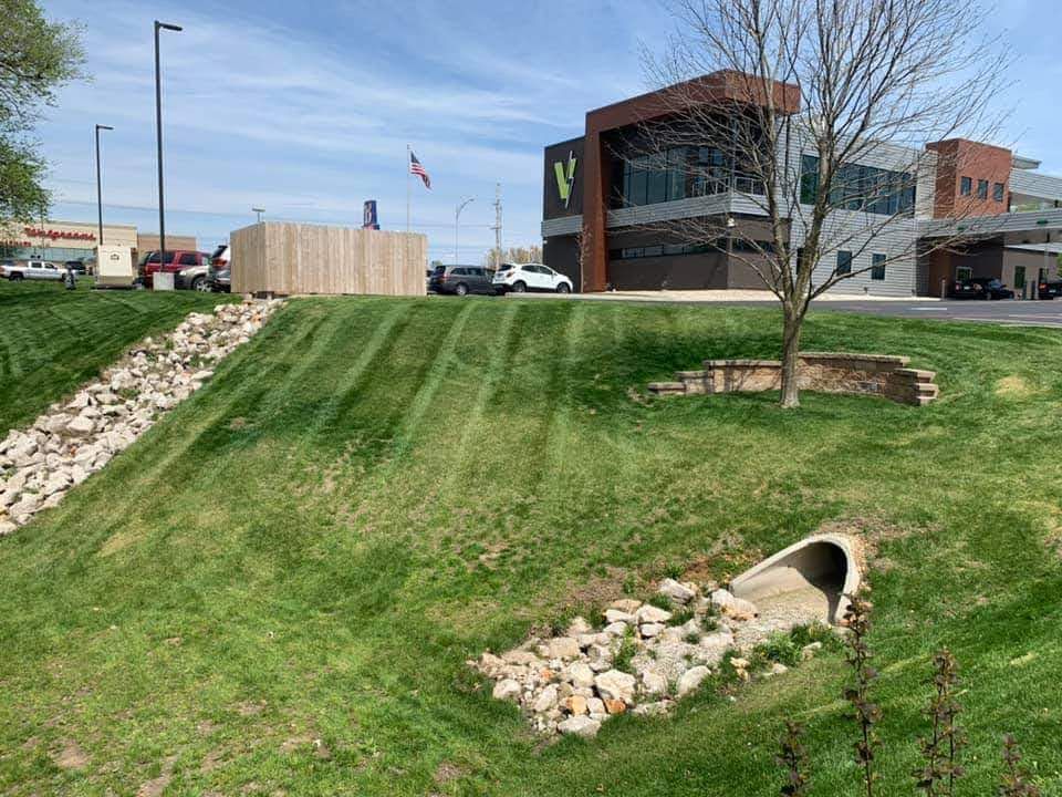 Grassy slope with drainage pipe, rocks, and building with V logo under blue sky.