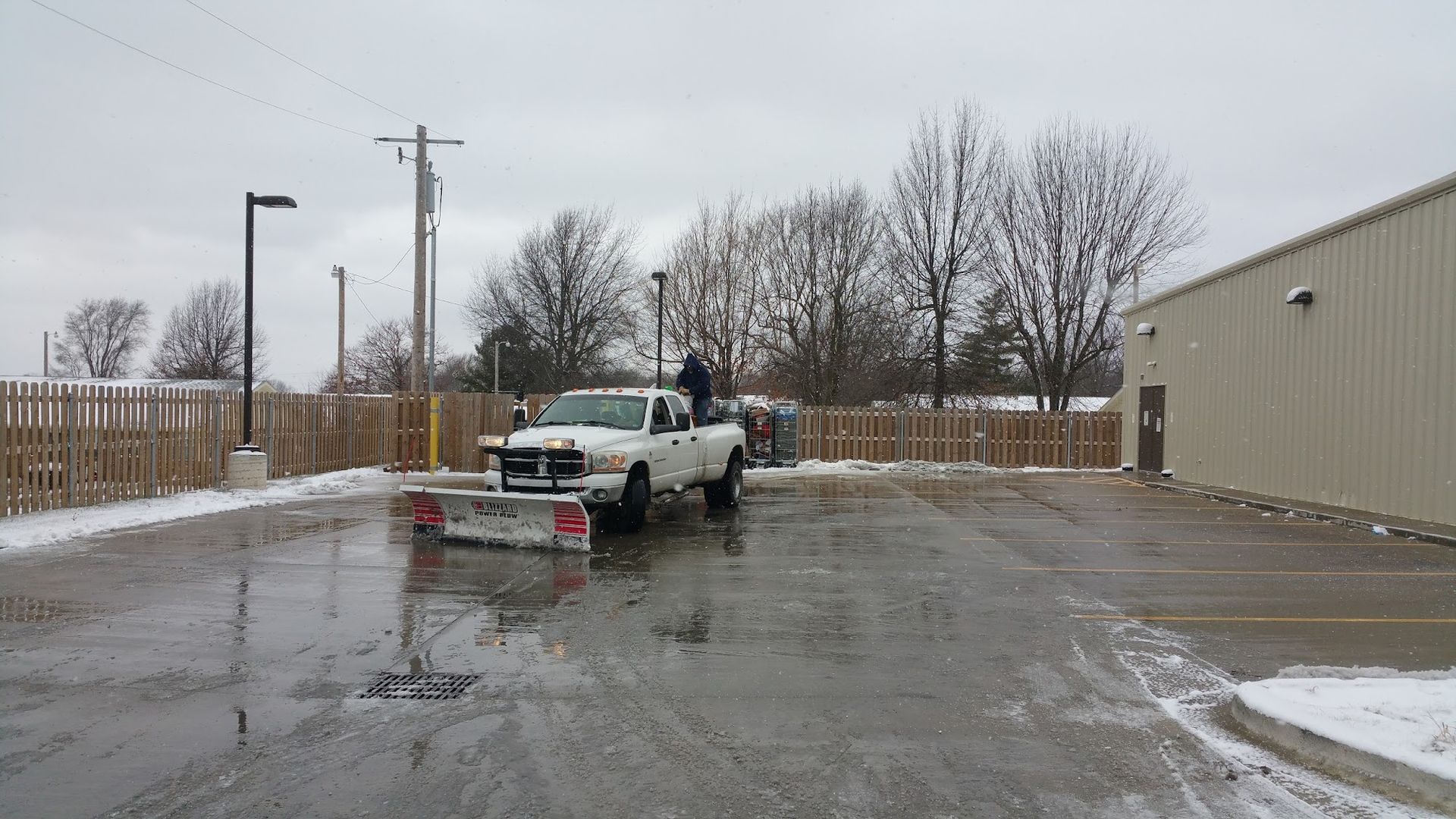 White pickup truck plowing snow in a parking lot, gray sky overhead.