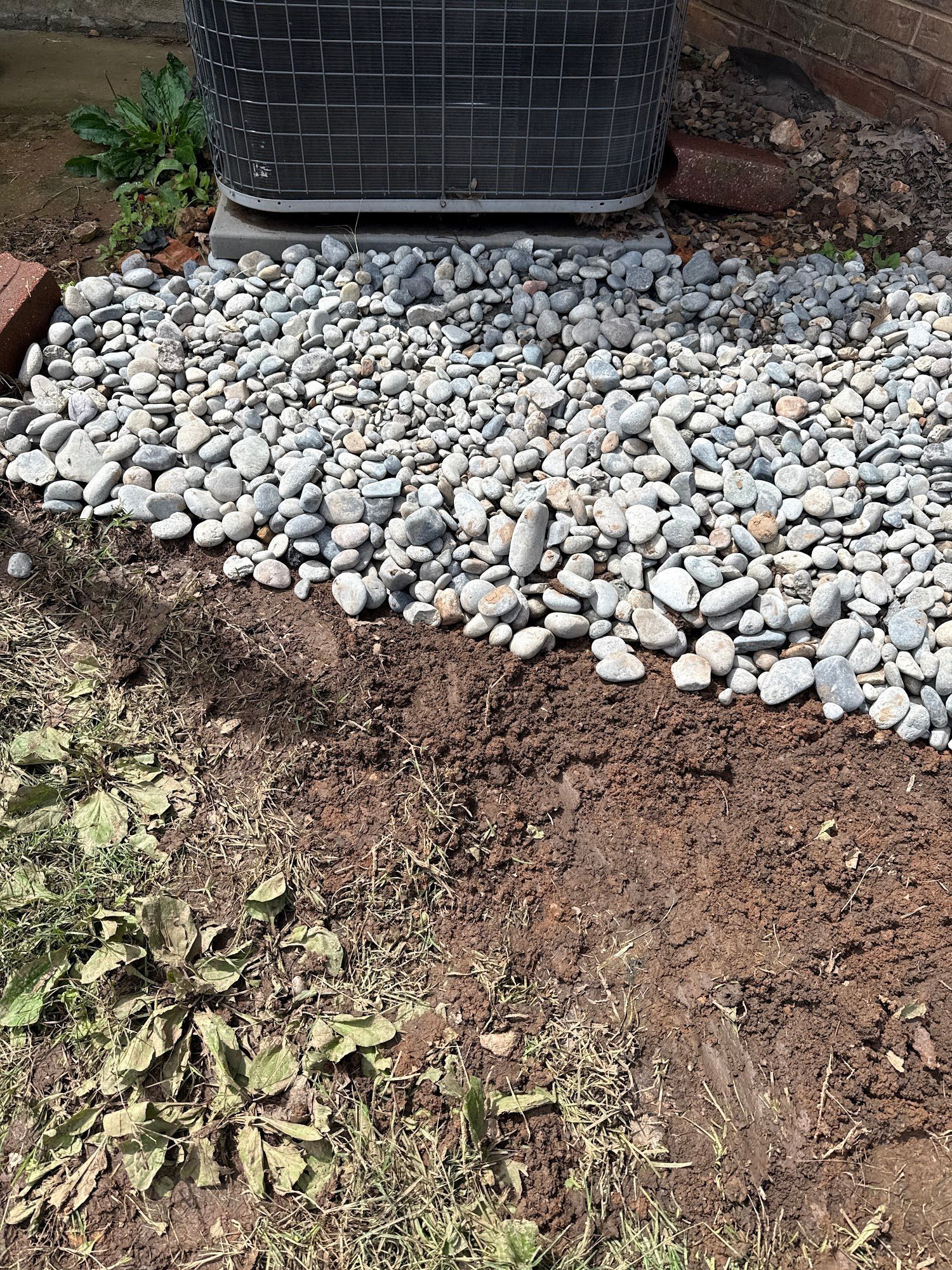 Gravel bed surrounds a gray air conditioner unit; adjacent dirt with sparse weeds.