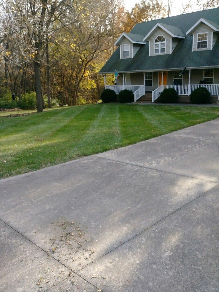 Green lawn with striped mowing pattern, house with green roof, and concrete driveway.