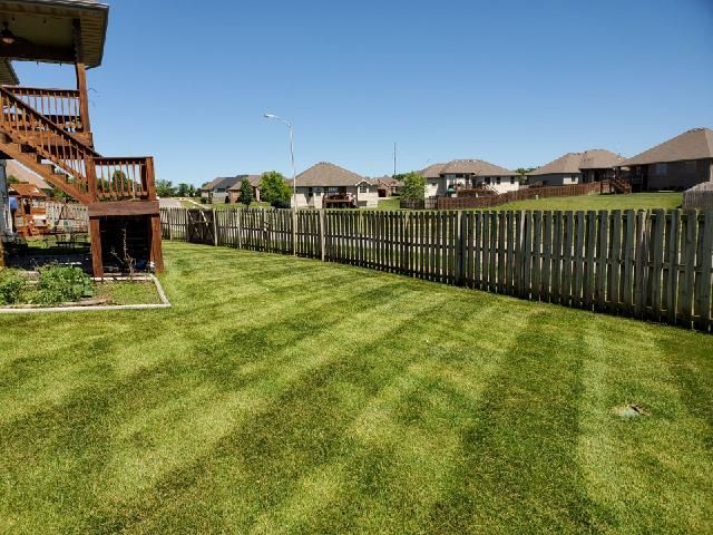 Lawn mowed with stripes, next to a wooden fence. Houses in the background under a blue sky.
