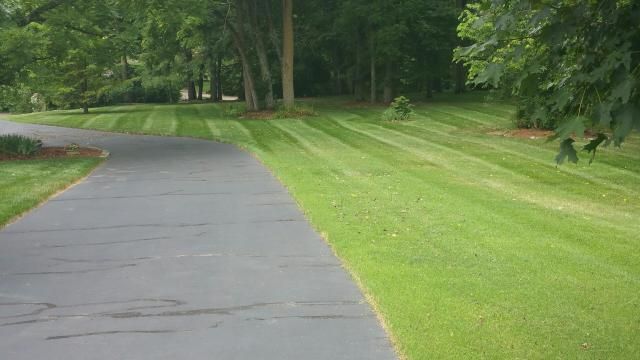 Asphalt driveway next to neatly striped green lawn, trees in background.