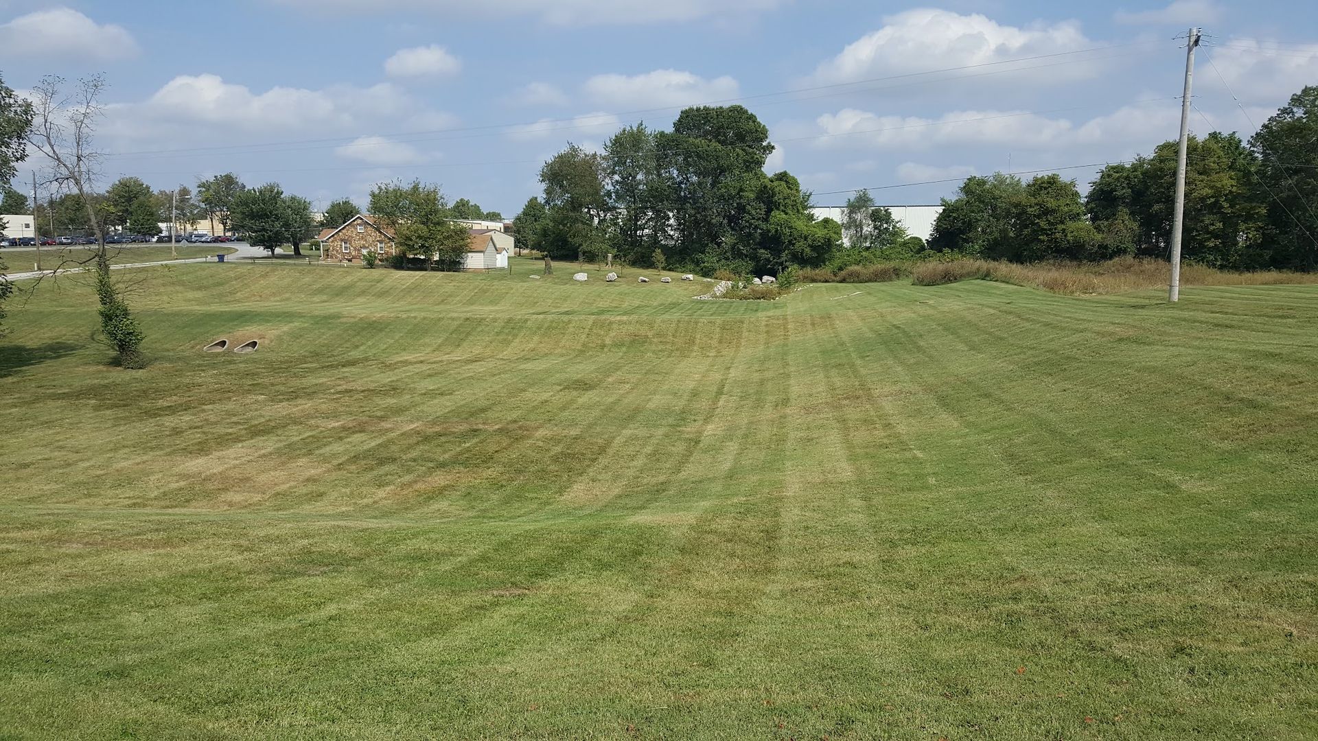 Mowed green field with a few trees and buildings in the background under a partly cloudy sky.