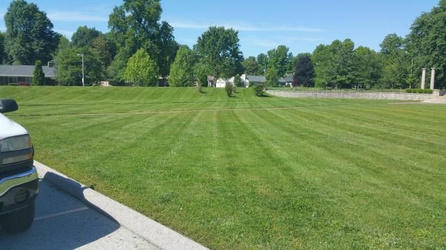 Green lawn with freshly cut stripes. Trees and buildings in the background. A white vehicle in the foreground.
