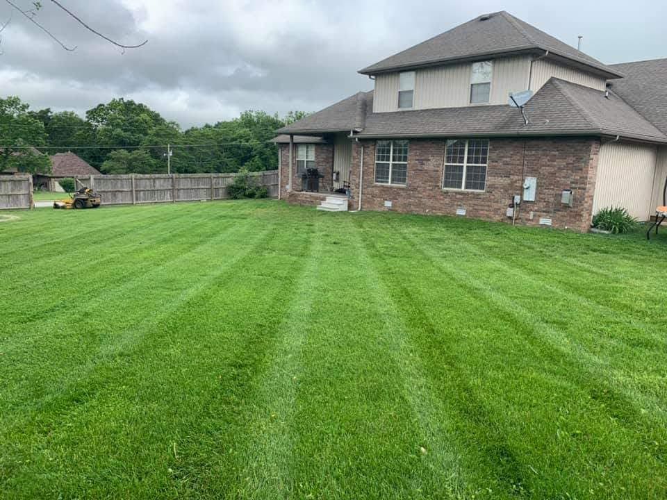 Green lawn with mowing stripes in front of a brick house and wooden fence on a cloudy day.