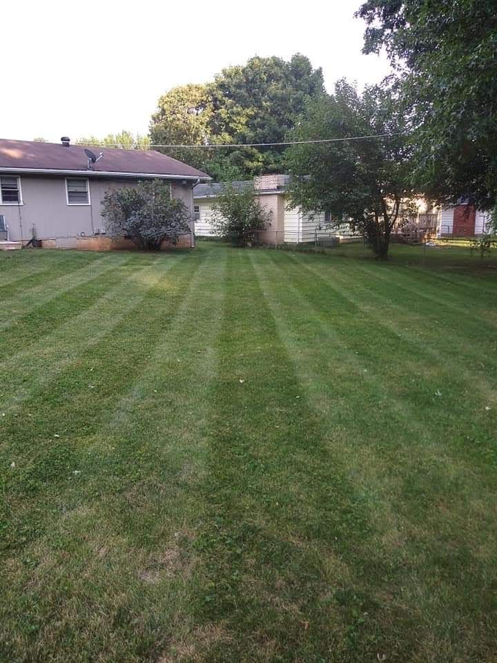 Lawn with striped pattern, mowed and green, with a house and trees in the background under a cloudy sky.