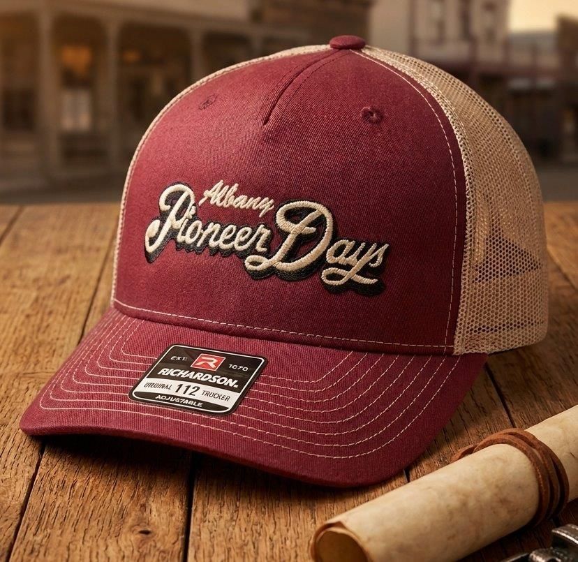 Maroon Albany Pioneer Days baseball cap with tan mesh back on a wooden table, beside a rolled parchment