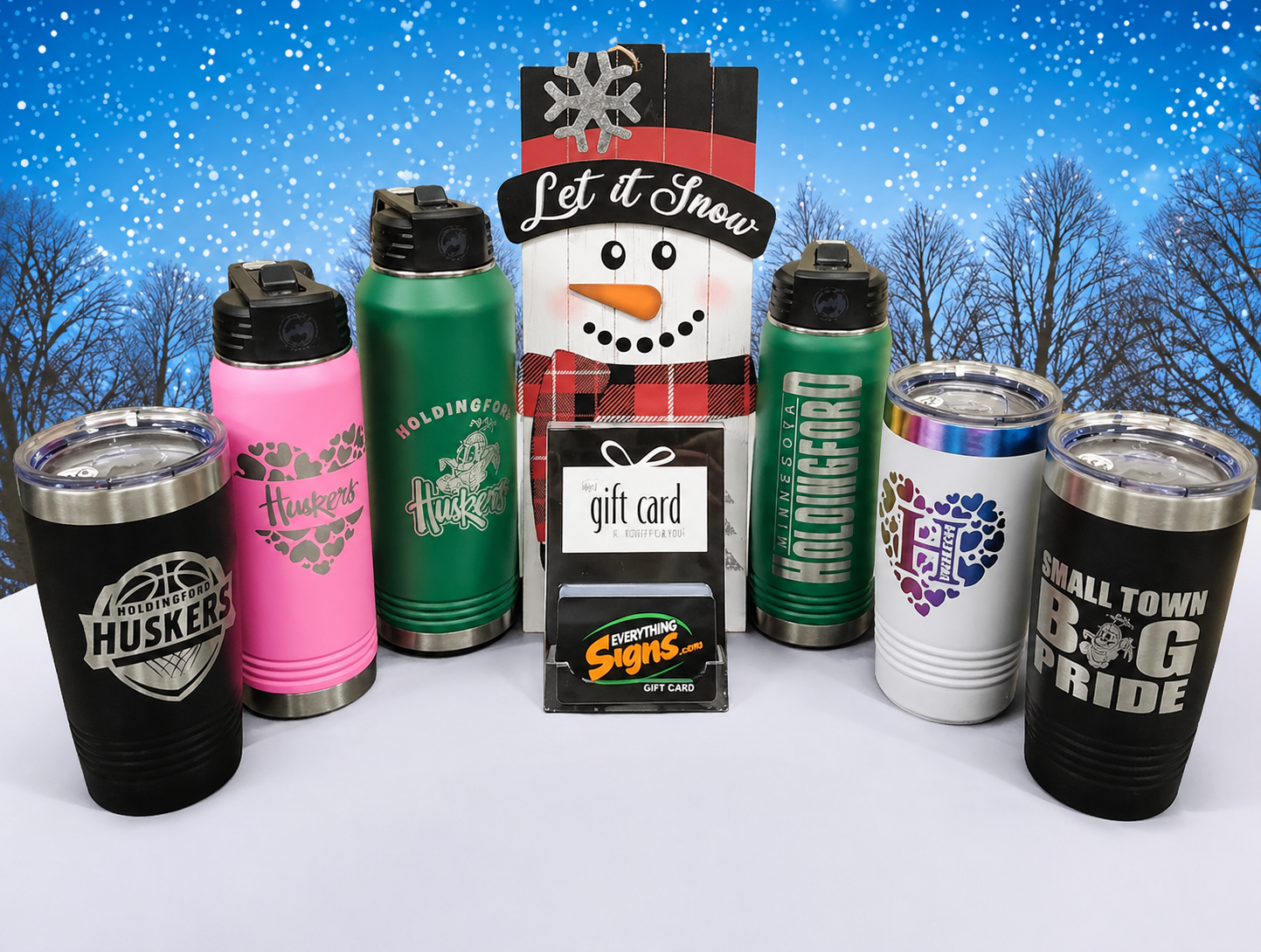 Assorted colorful tumblers and mugs arranged in snow, with a snowman-themed gift box in the center.