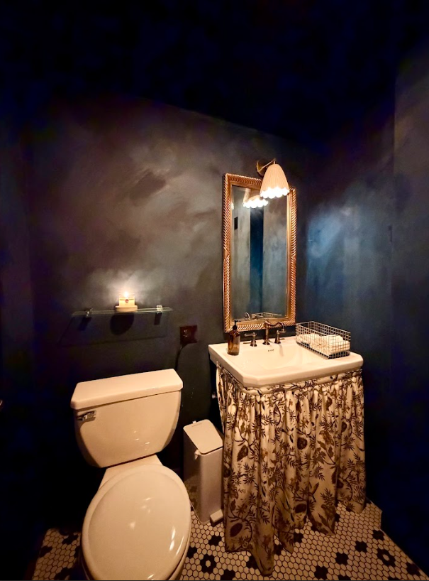 Dim bathroom with navy walls, white toilet, sink, ornate mirror, and patterned black-and-white tile floor.