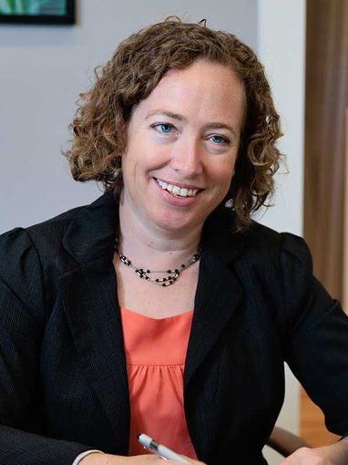 A woman in a black jacket and white shirt is smiling with her arms crossed
