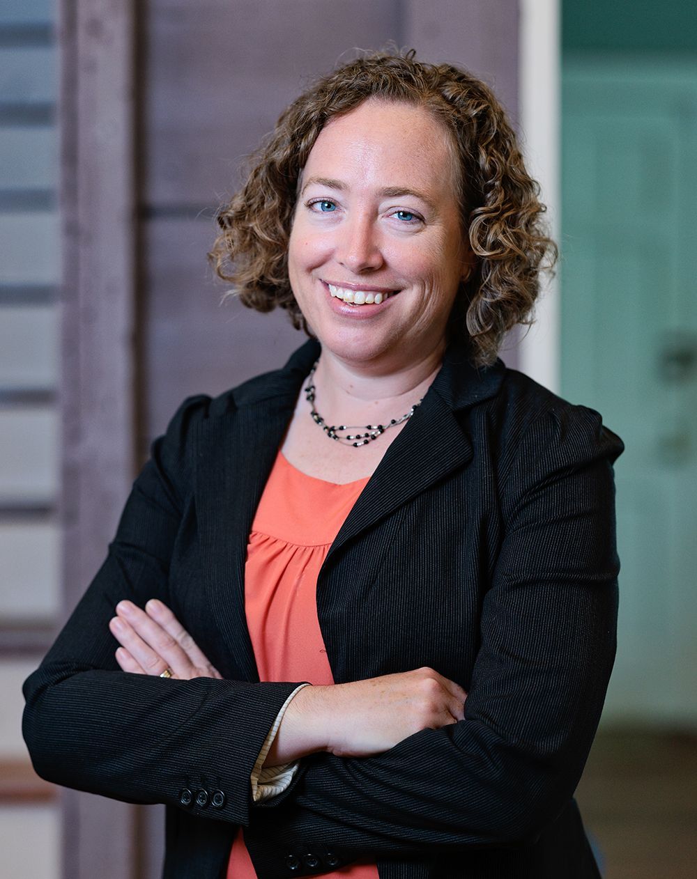 A woman in a black jacket and orange shirt is smiling with her arms crossed.