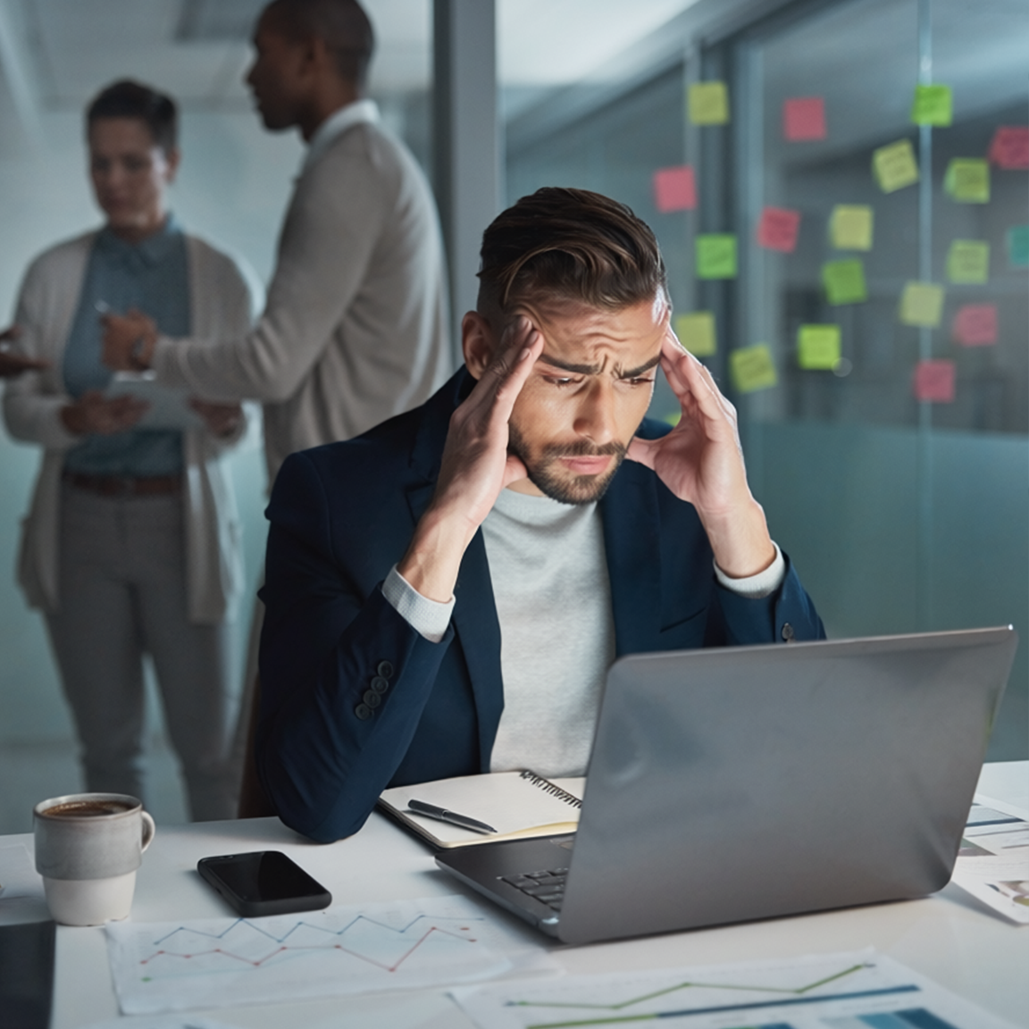Stressed man at laptop in a meeting room, holding his temples while coworkers talk in the background.