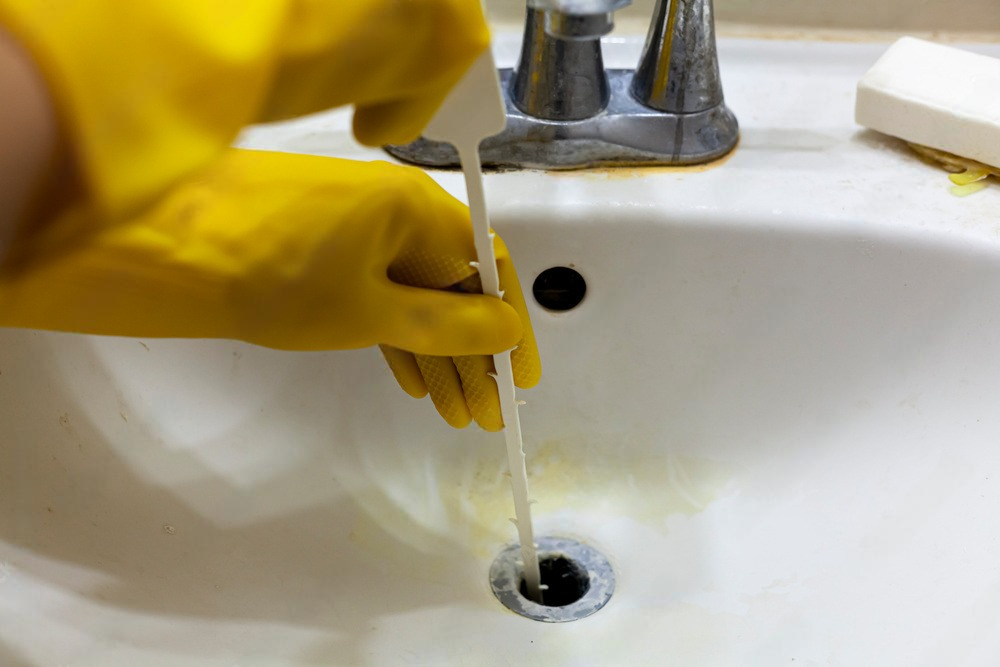 A person wearing yellow rubber gloves is cleaning a sink with a plunger.