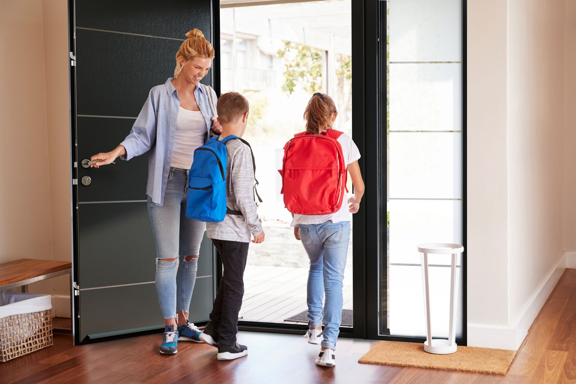 A woman is opening a door for two children with backpacks.