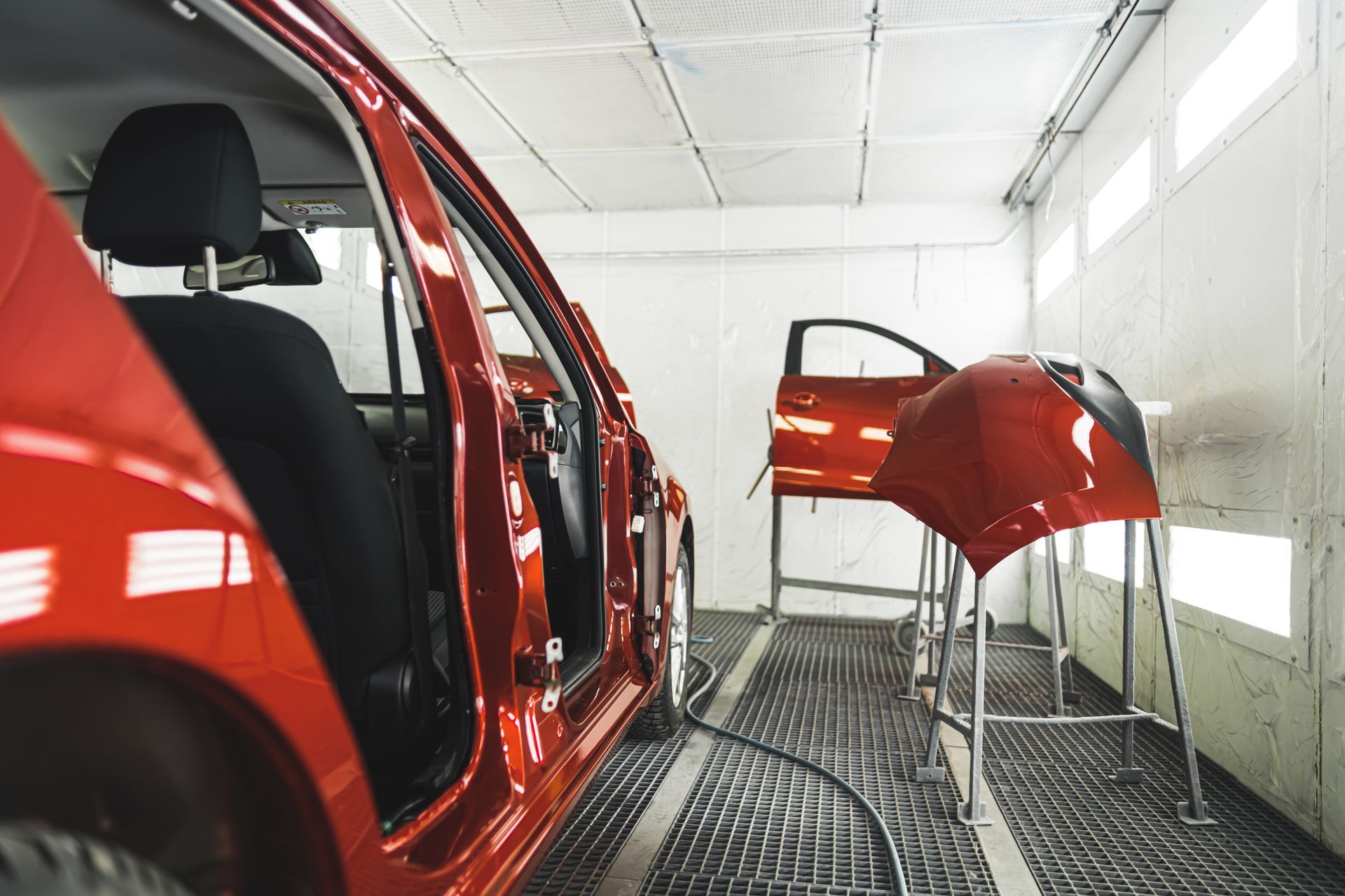 Red car in a paint booth with door and bumper on stands.