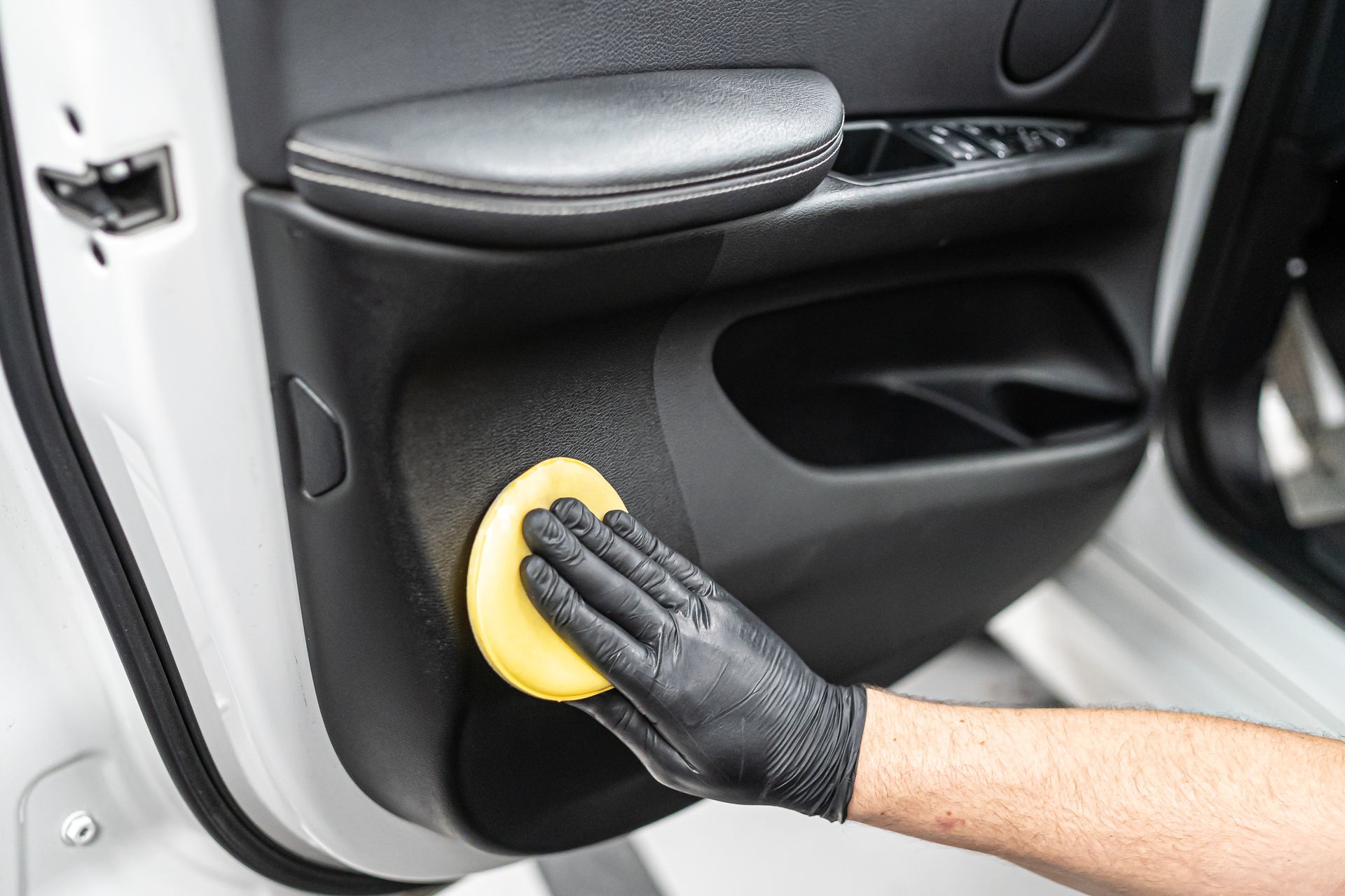Gloved hand applying a cleaning product to a car door panel with a yellow applicator. The panel is black.