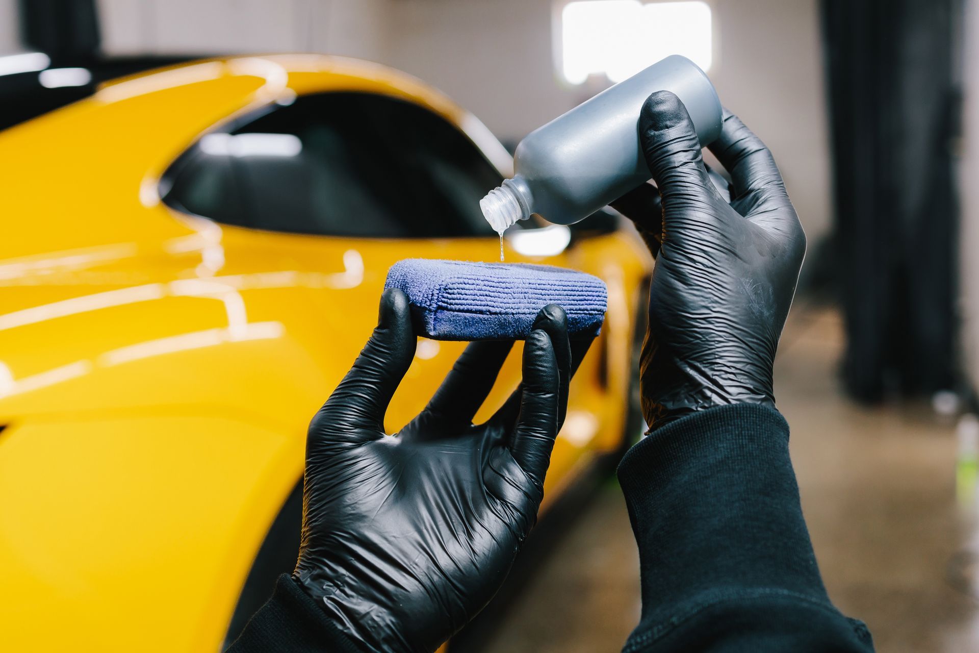 Hands in black gloves applying liquid product to blue applicator on a yellow car.