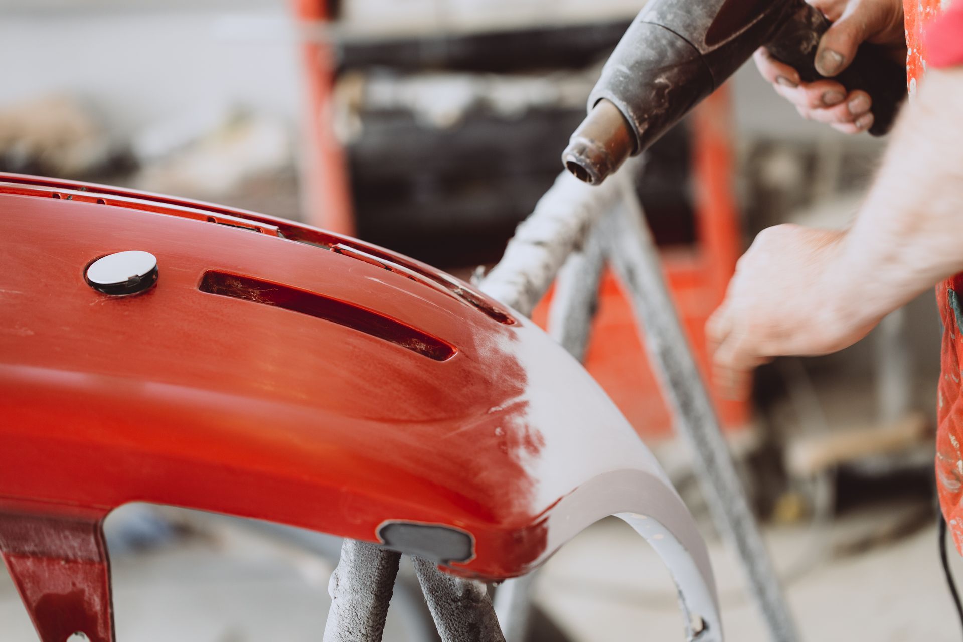 Person using a heat gun on a red car bumper for auto body repair.