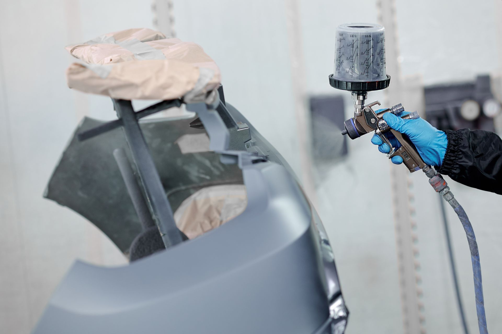 Person wearing gloves spray-painting a gray car bumper in a paint booth.