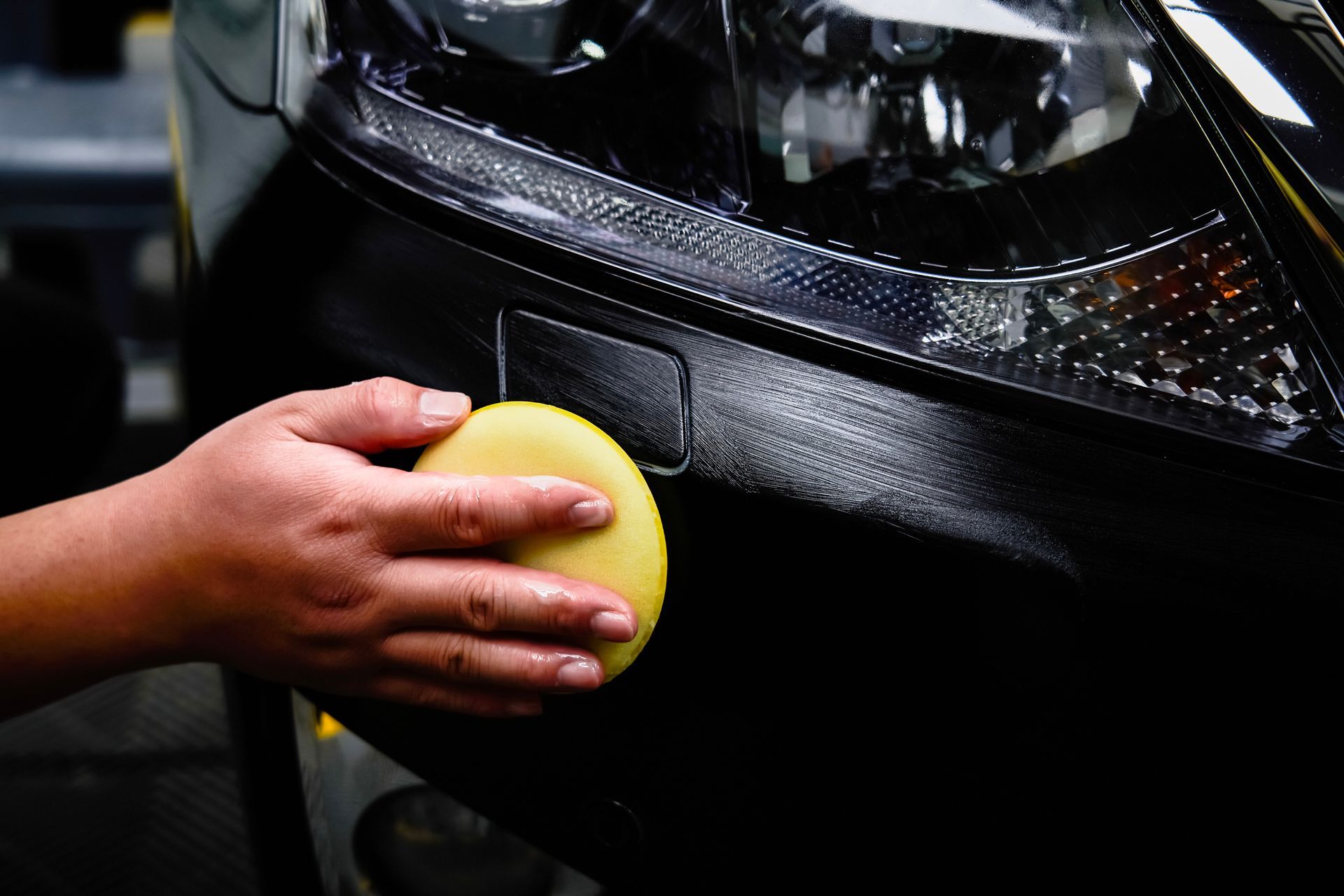 Hand using a yellow sponge to apply something to a black car bumper, near a headlight.