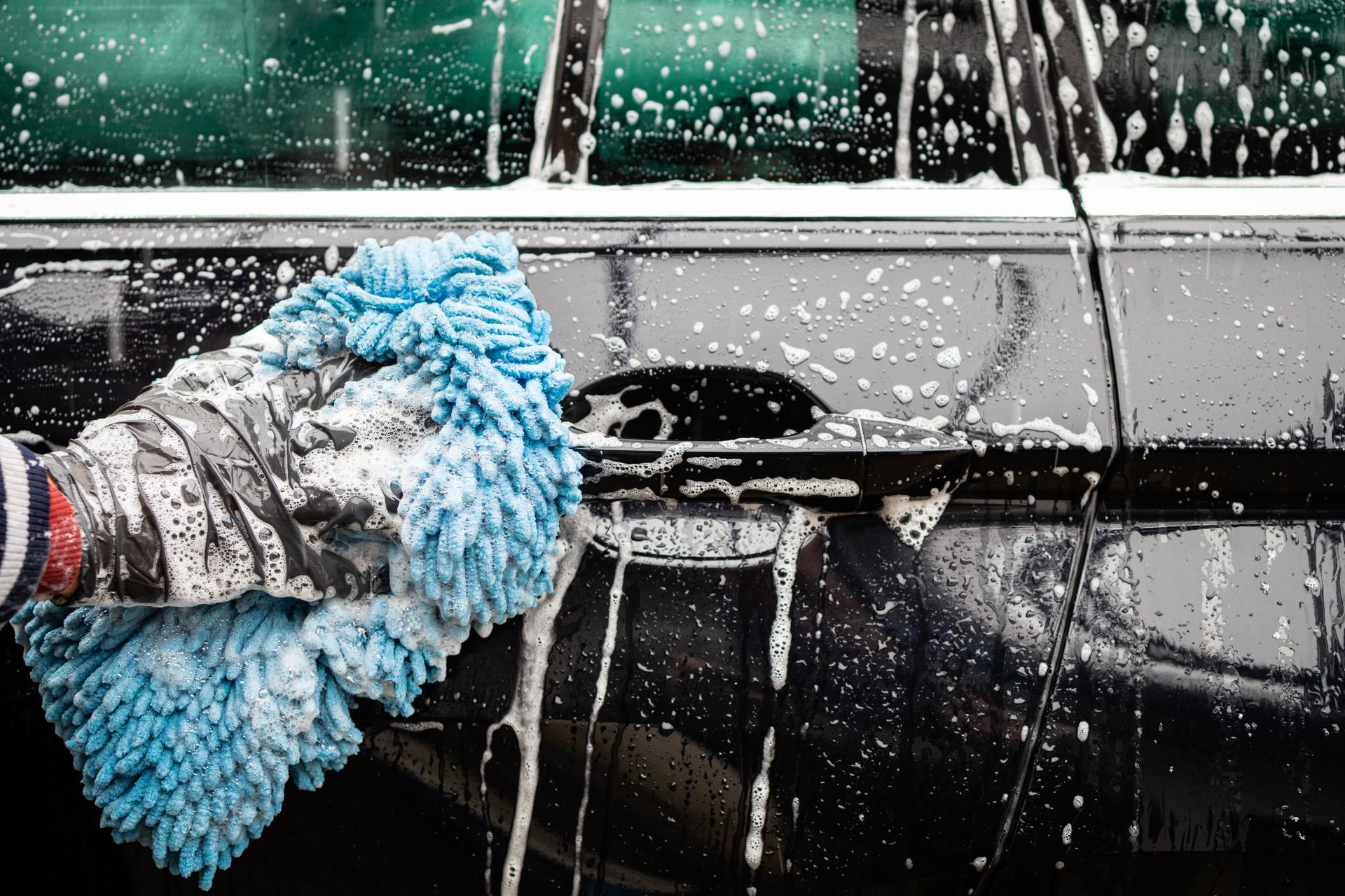 A gloved hand washes a black car with a blue microfiber mitt, covered in soap suds.