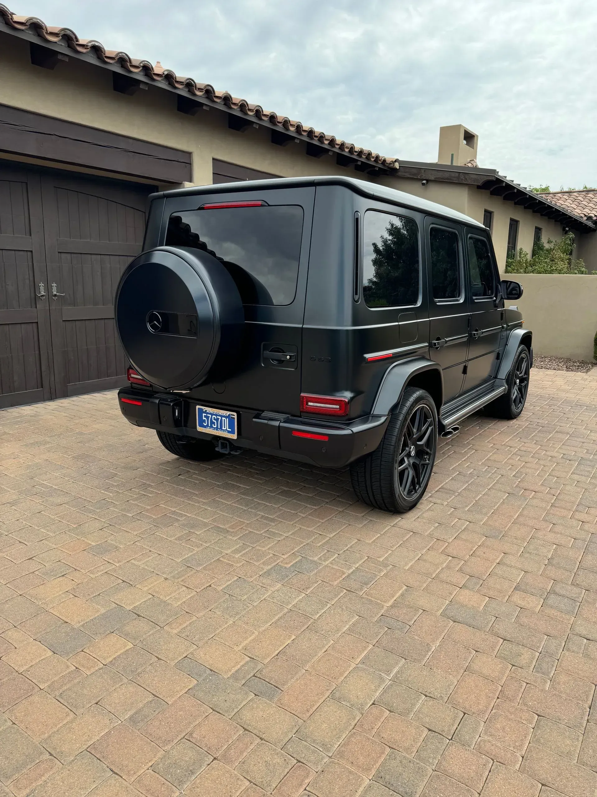 Black Mercedes-Benz G-Class SUV parked on a brick driveway, near a beige building with a brown garage door.