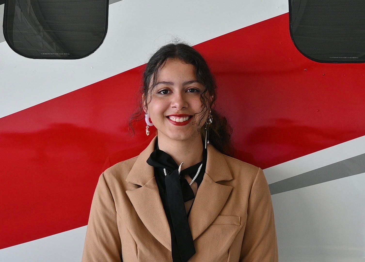 A woman in a tan jacket and black tie is standing in front of a red and white wall.