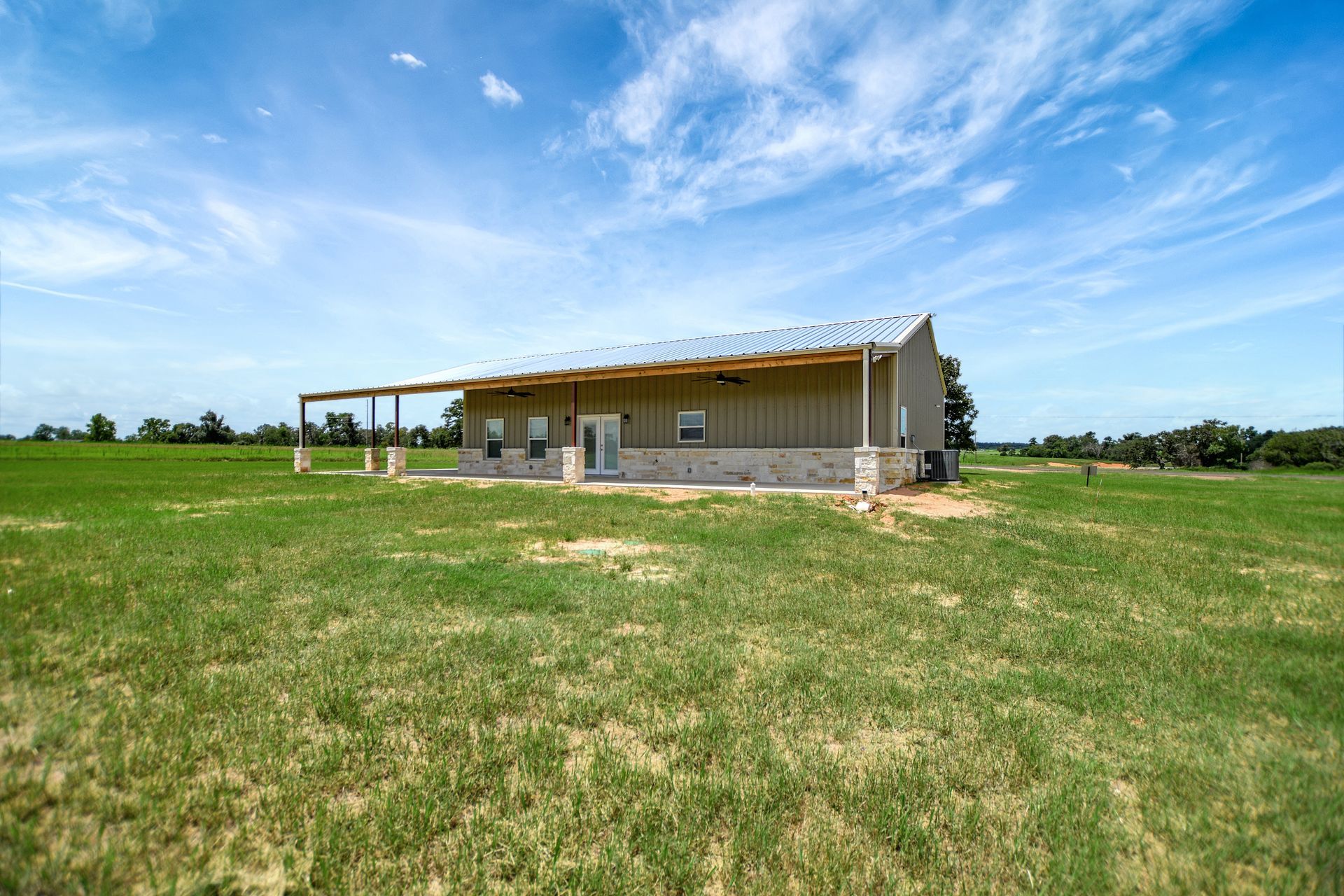 A modern, tan metal building with a covered porch in a grassy field under a blue sky.