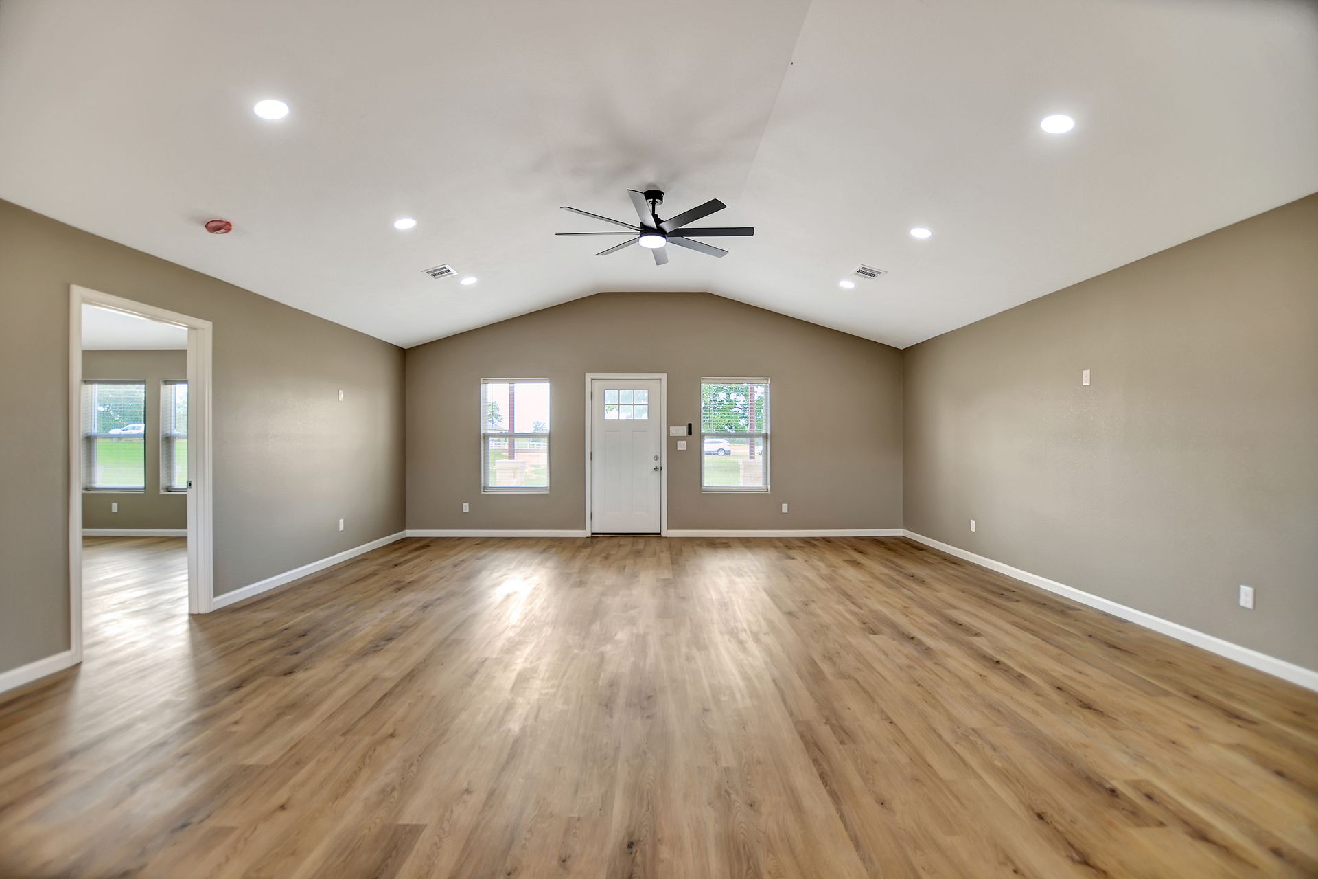 Empty living room with wood floors, tan walls, and a vaulted ceiling with a fan.