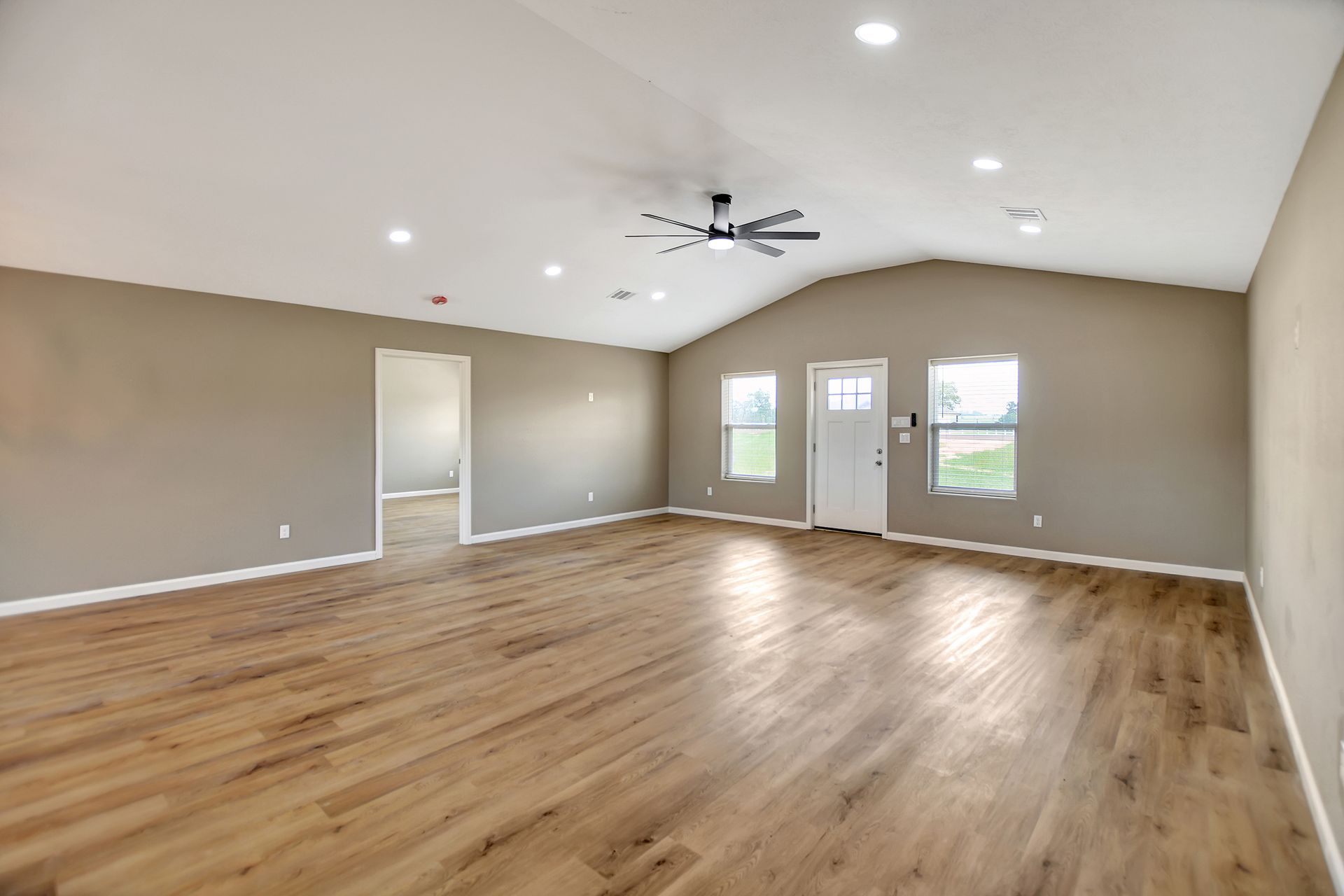 Empty, open-concept living room with wood floors, beige walls, high ceiling, and natural light.