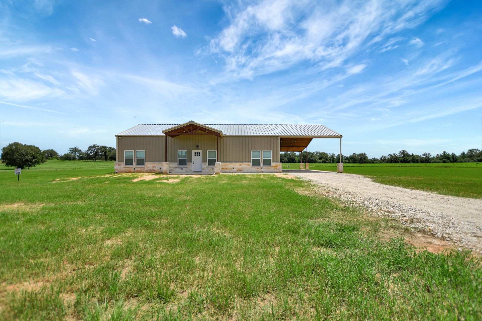 Tan metal home with covered porch, surrounded by green field under blue sky.