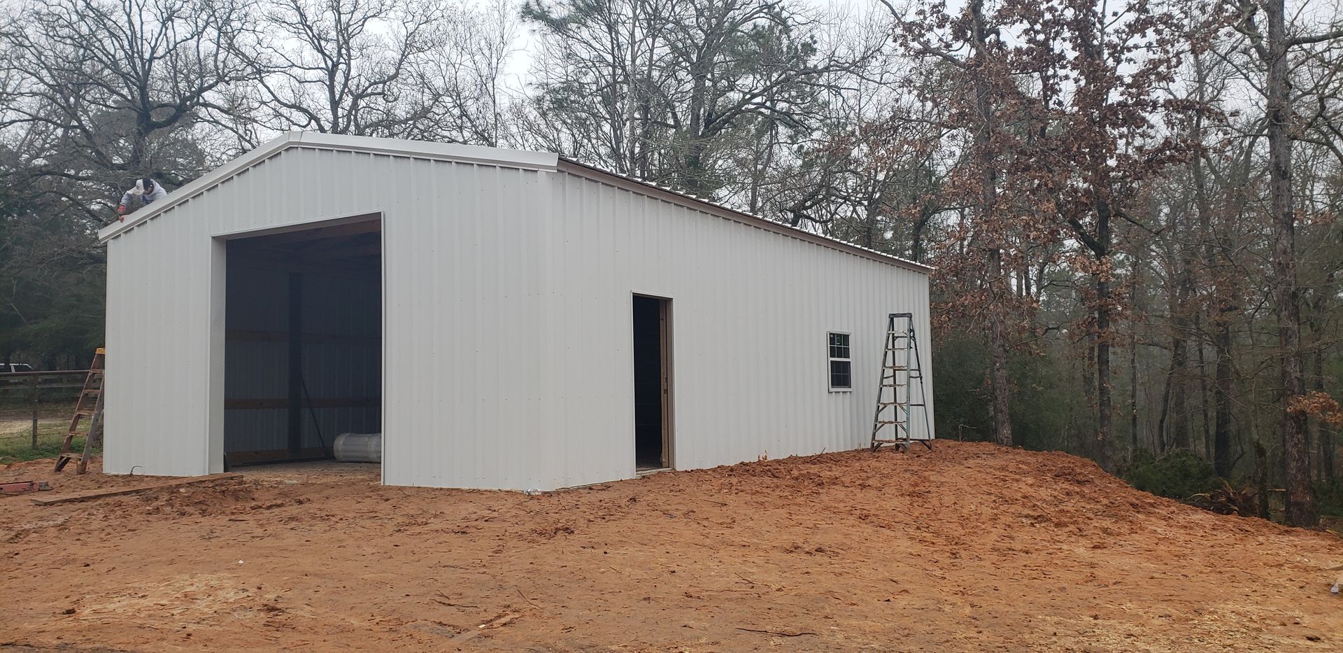 A white metal building with a large opening, door, and window stands on a dirt mound; trees in the background.