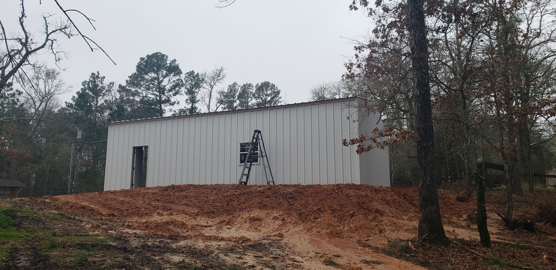 A white metal building sits on a dirt foundation, surrounded by trees. A ladder leans against the building.