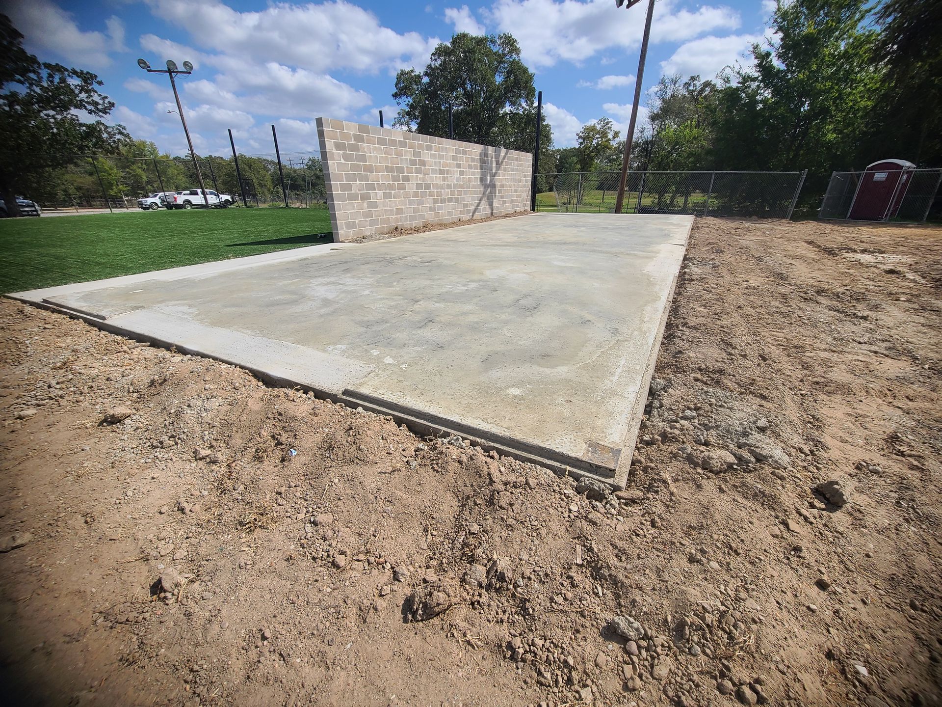 Concrete slab in dirt, partially completed brick wall, grassy area, blue sky.