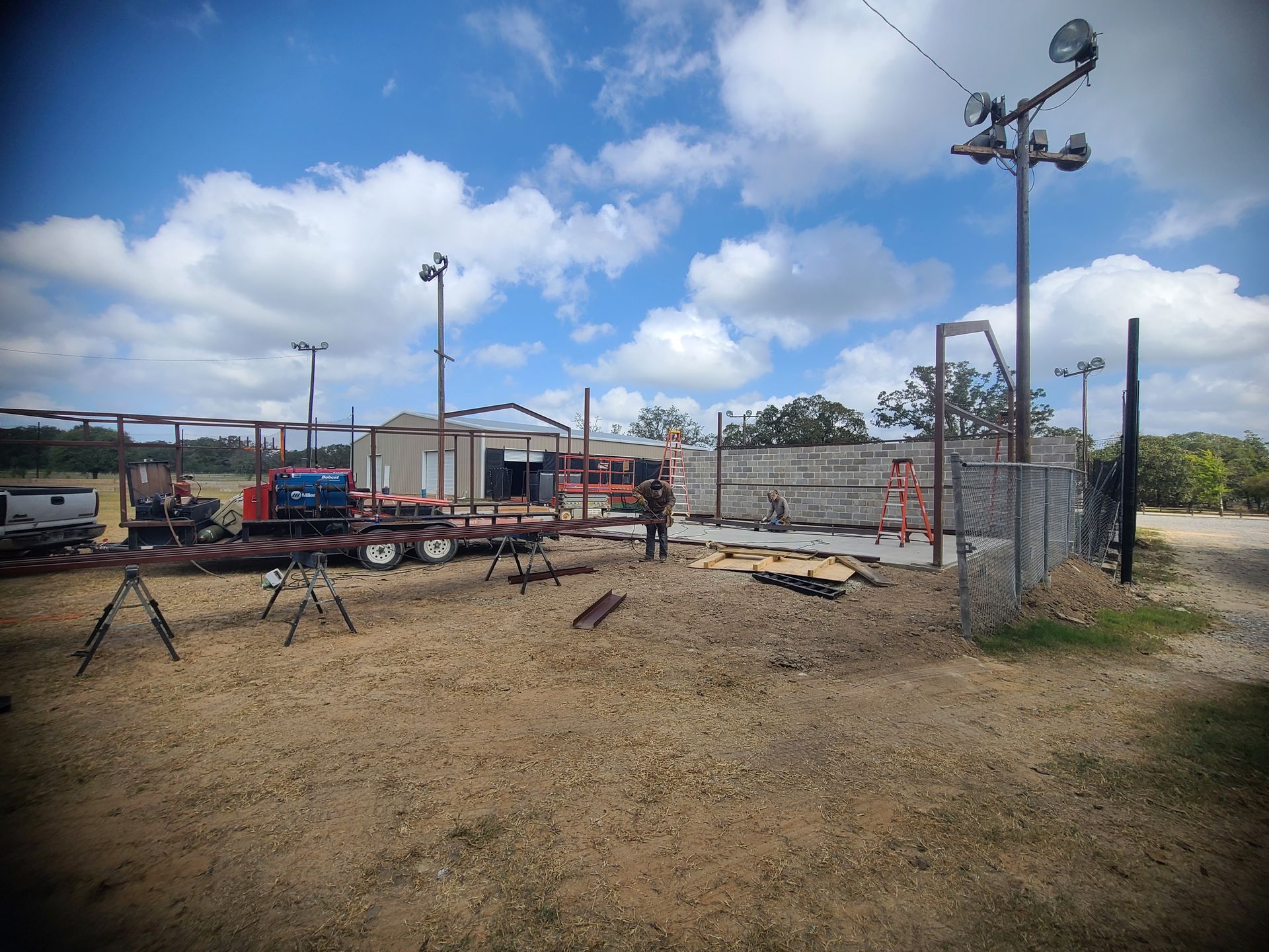 Oil field with equipment, metal pipes, a trailer, and person standing outdoors under a cloudy sky.