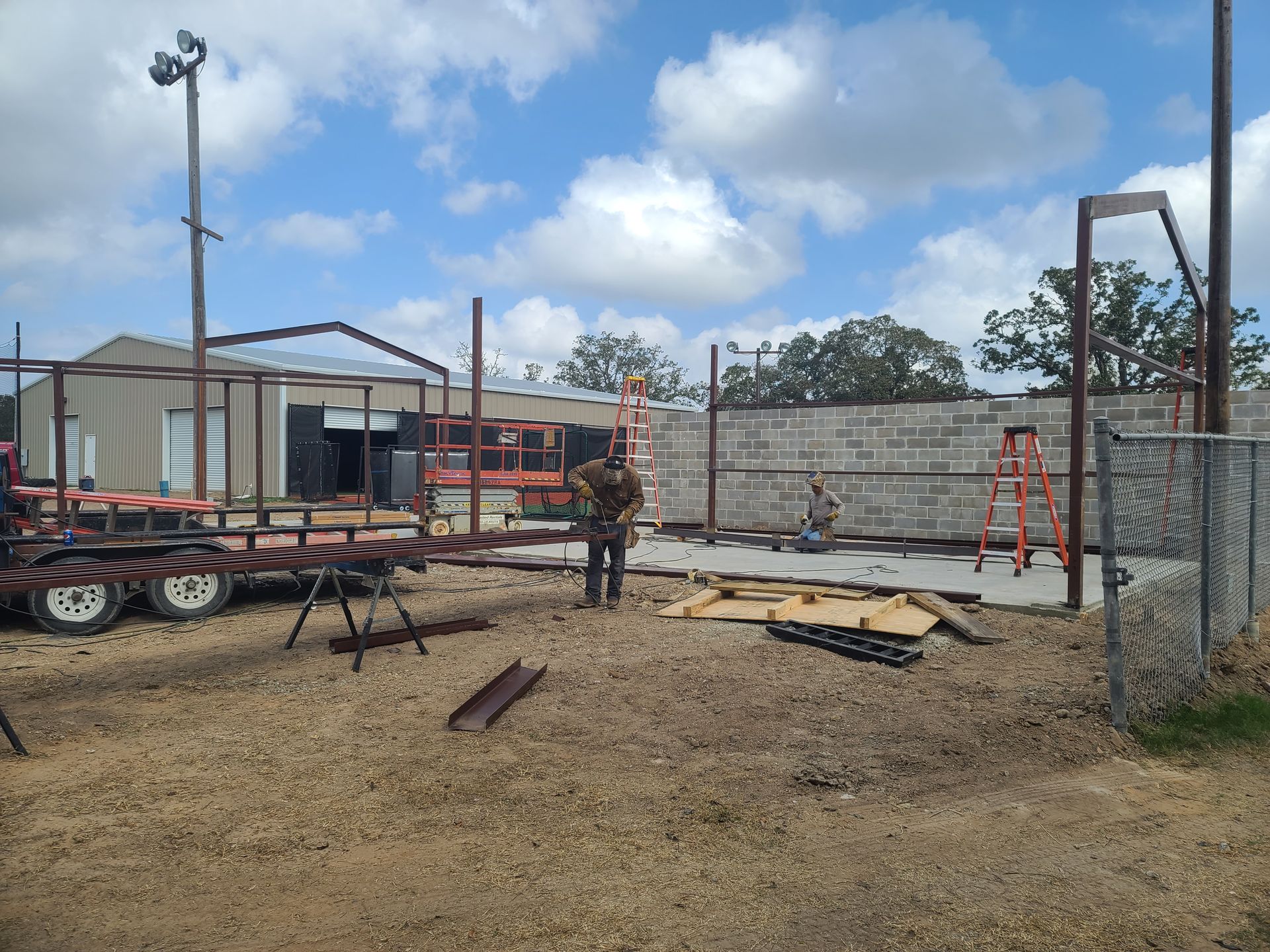 Construction site with workers; metal framework, trailer, cinder block wall. Blue sky, overcast.