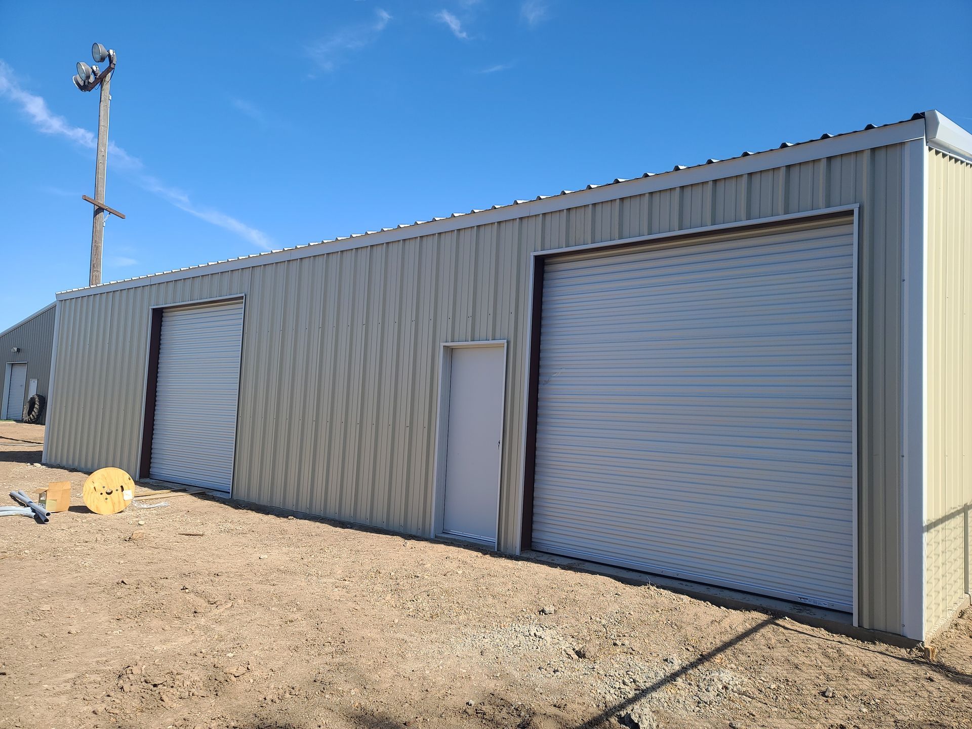 Tan metal building with two garage doors, a doorway, and a clear blue sky.