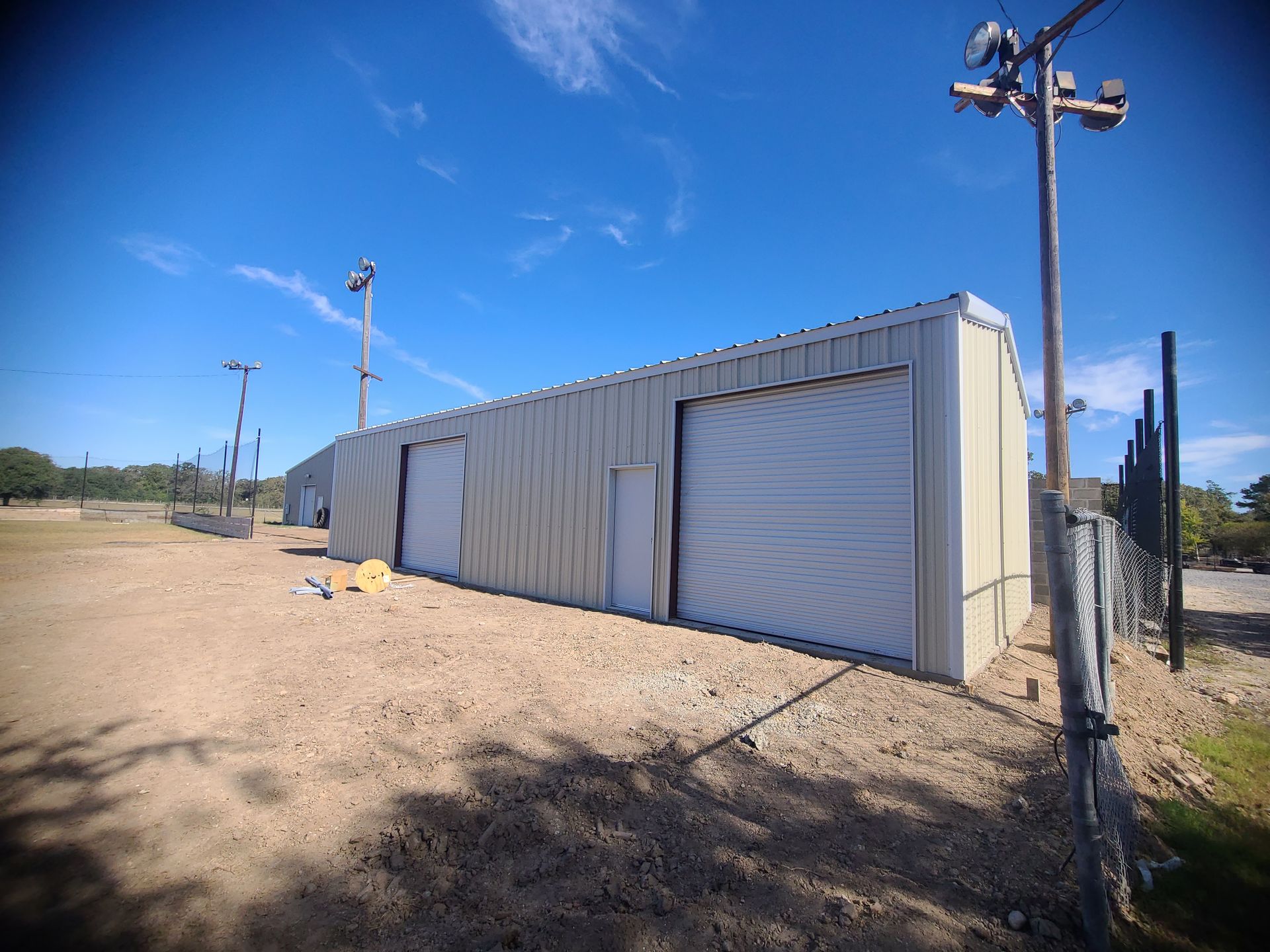 A tan metal building with two doors sits on a gravel lot under a blue sky, with utility poles nearby.