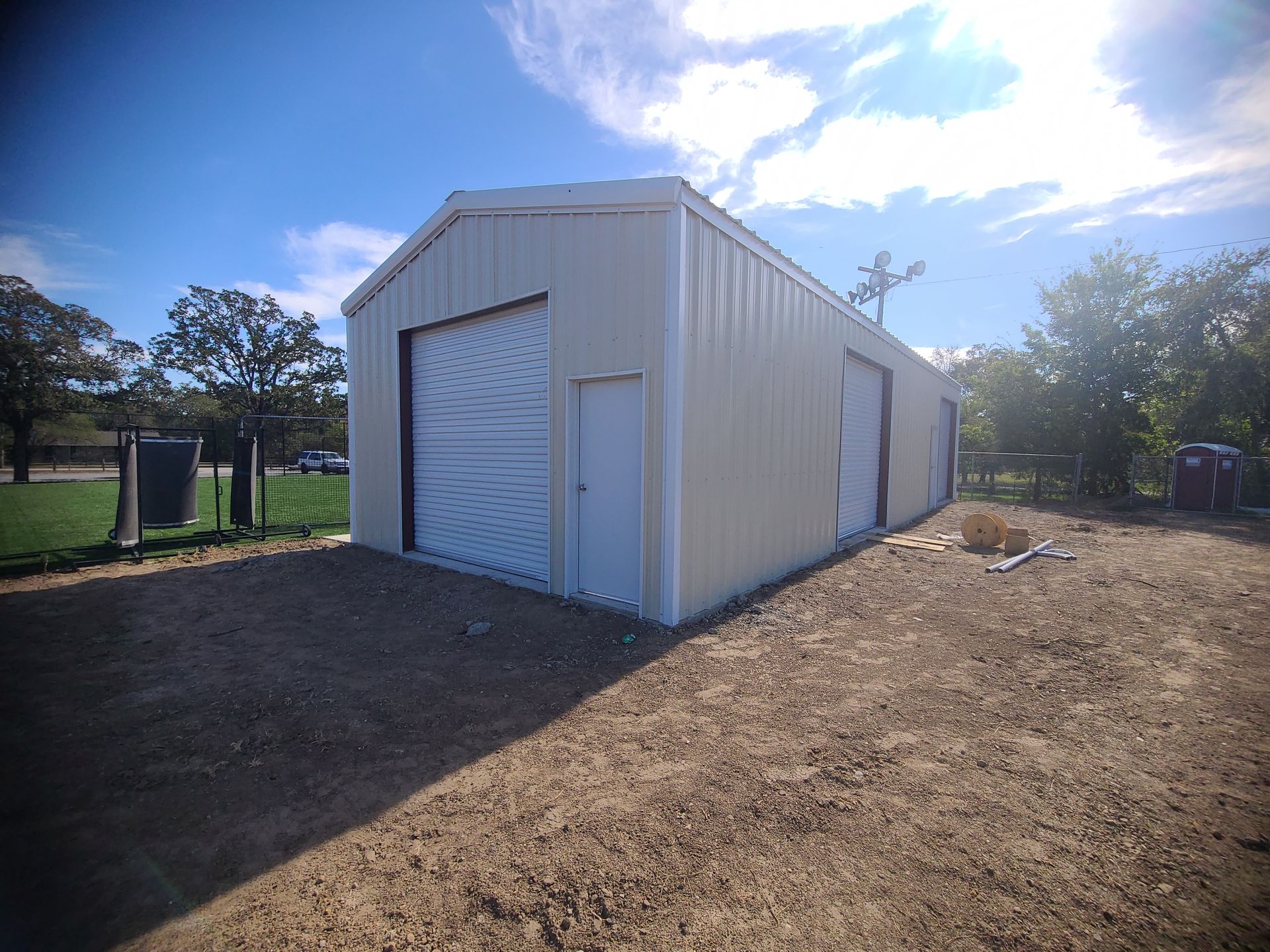 Tan metal building with three bays, two garage doors and a man door on gravel, sunny day.