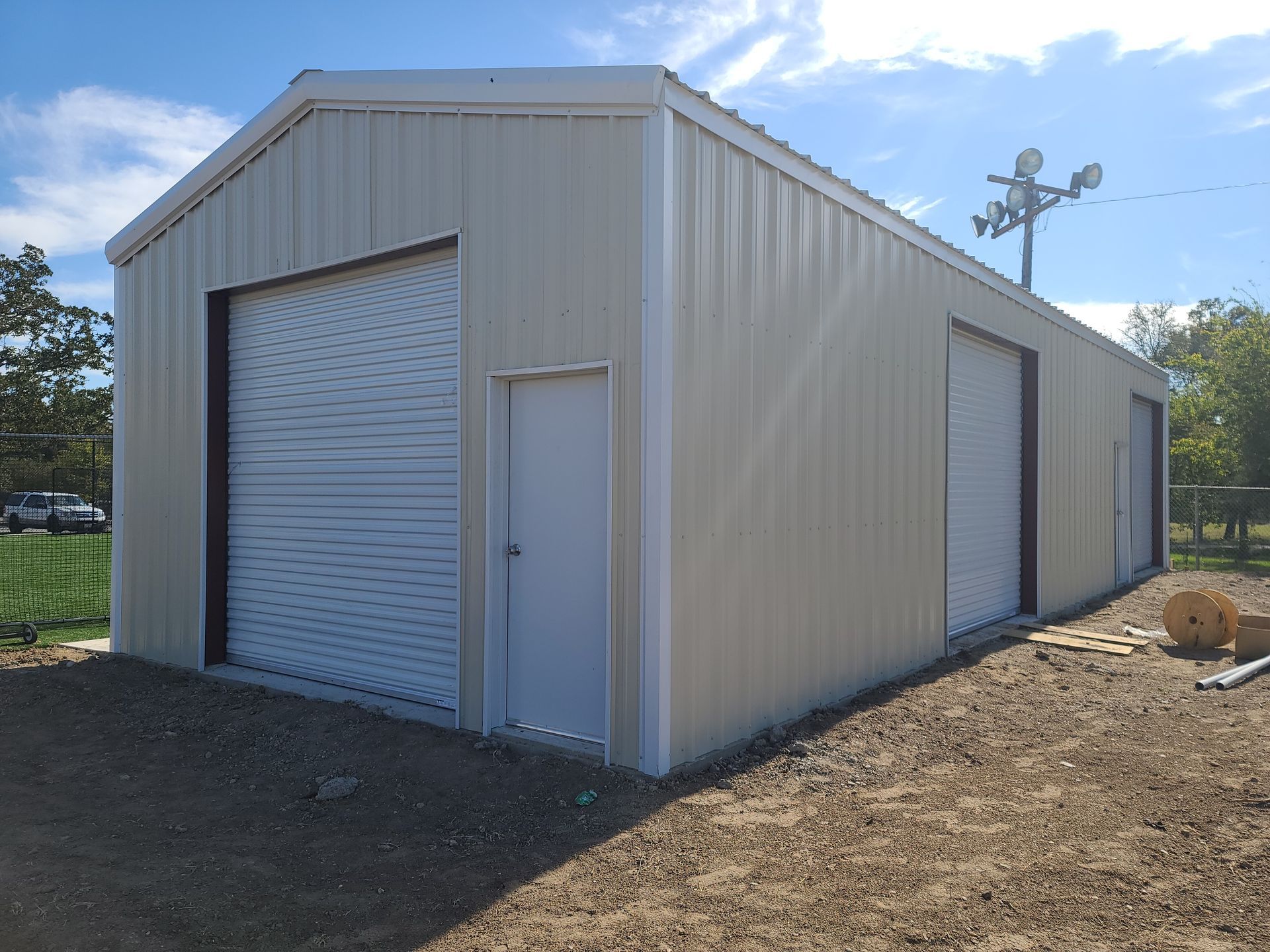 Tan metal building with two garage doors and a side door, on a gravel lot under a blue sky.