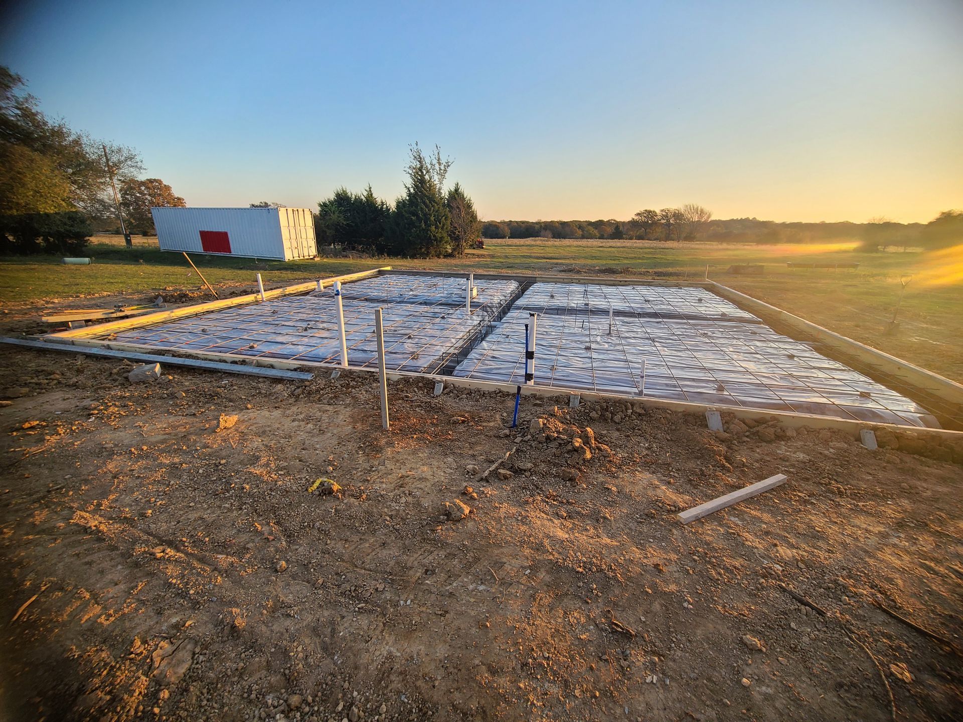 A dirt plot with gray gravel and pipes enclosed by wooden frames; background includes a white building and green fields.
