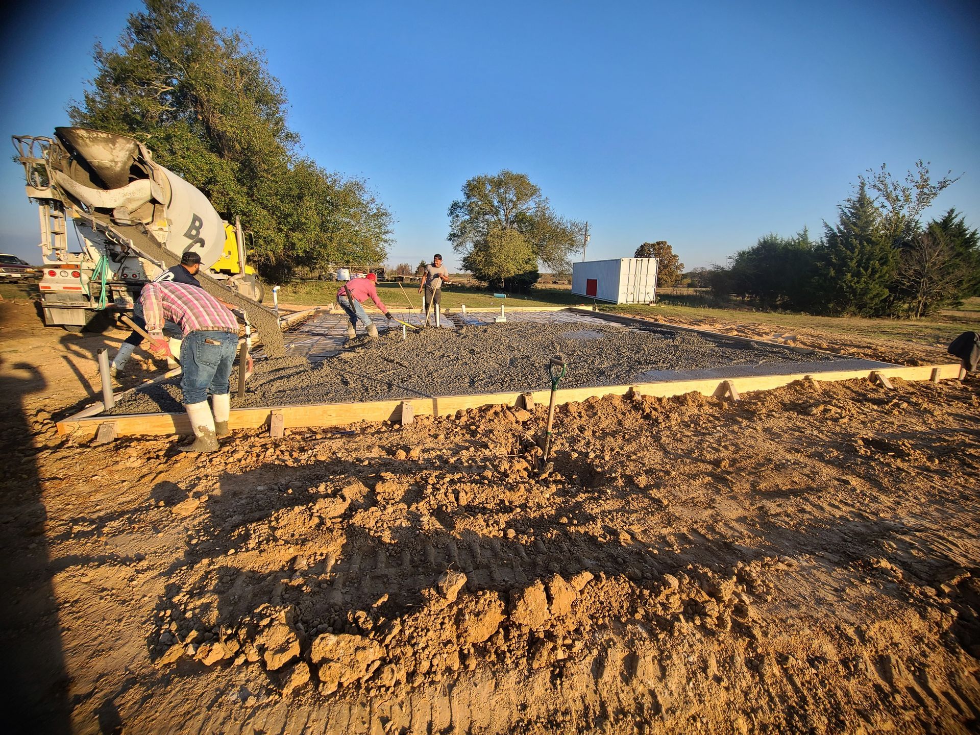 Concrete being poured into a wooden frame by workers with a cement truck in a dirt field.