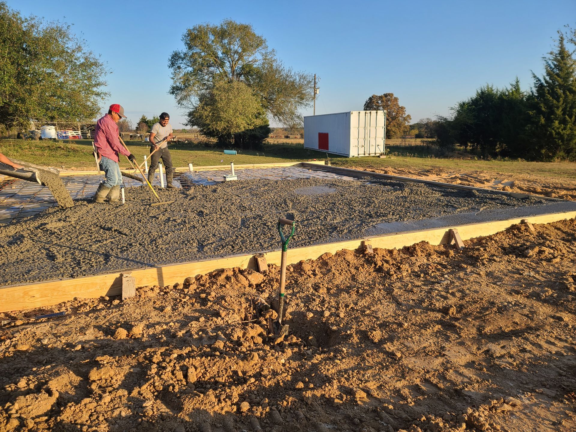 Workers pouring and leveling concrete for a foundation in a grassy outdoor area.