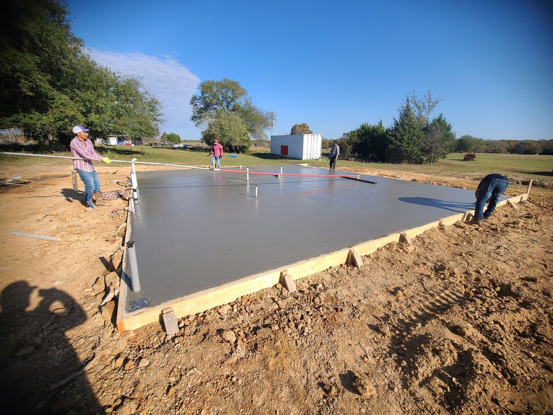 Workers smoothing fresh concrete slab for a building foundation. Brown earth, blue sky, sunny day.
