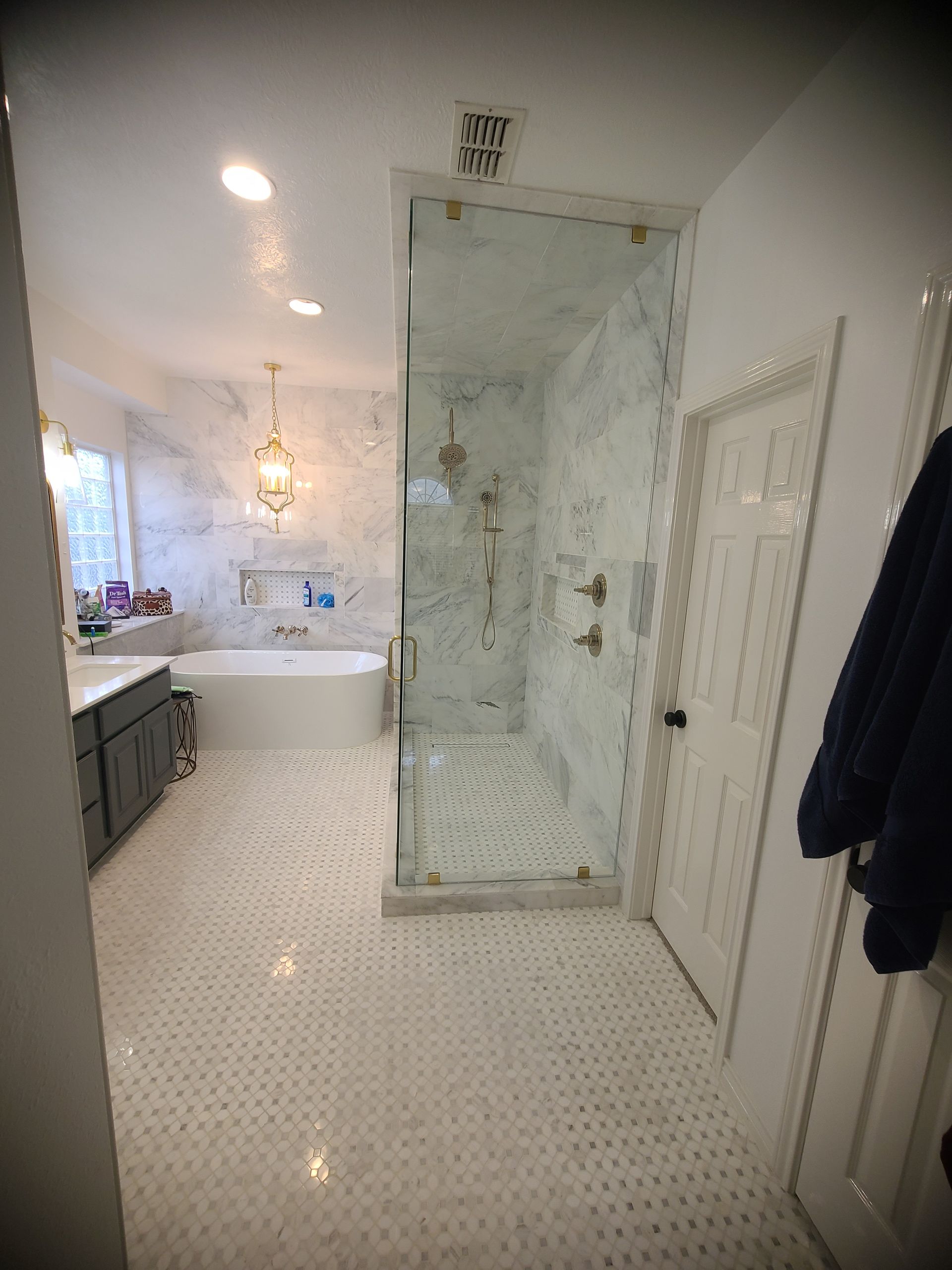 A spacious bathroom with a glass shower, marble walls, a white bathtub, and gray cabinets.