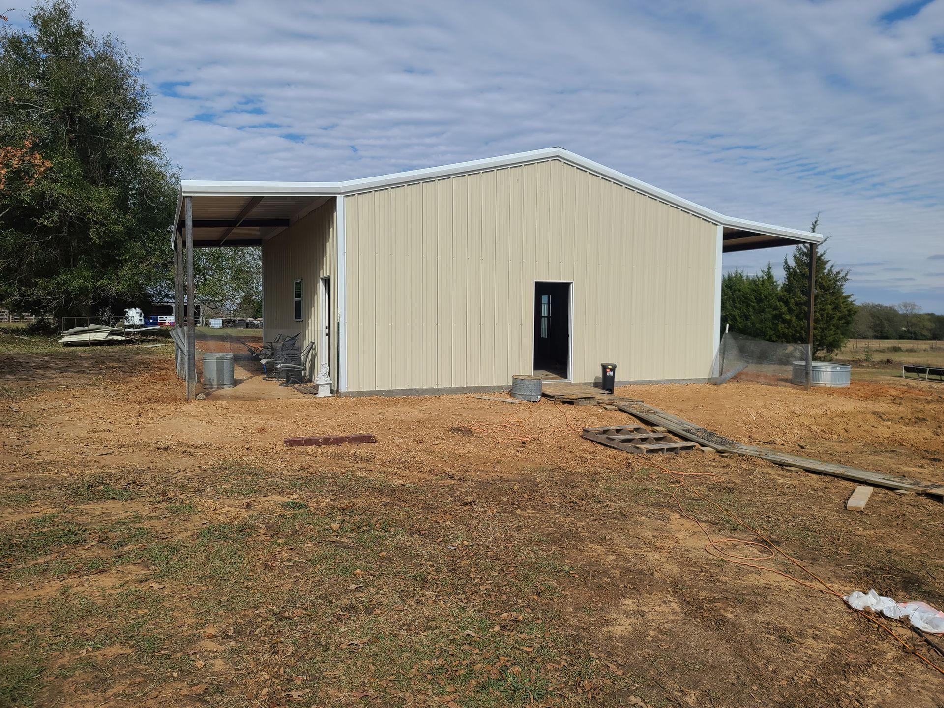 Beige metal building with a covered porch in a grassy field under a cloudy sky.