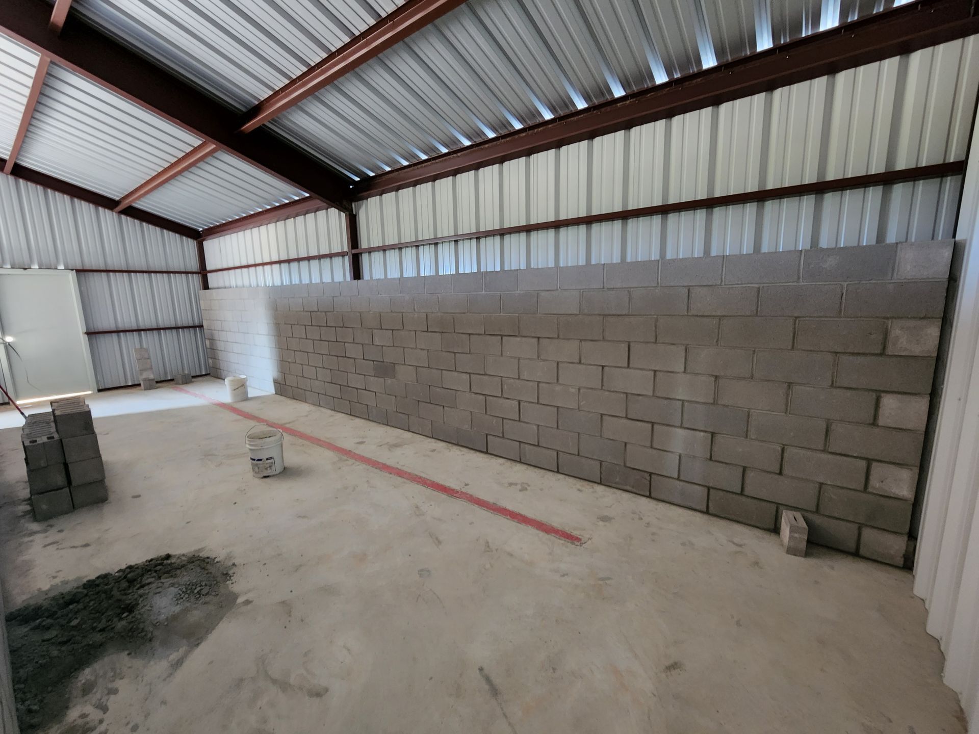 Inside a metal-roofed building under construction; cinder block wall is being built, with concrete floor.