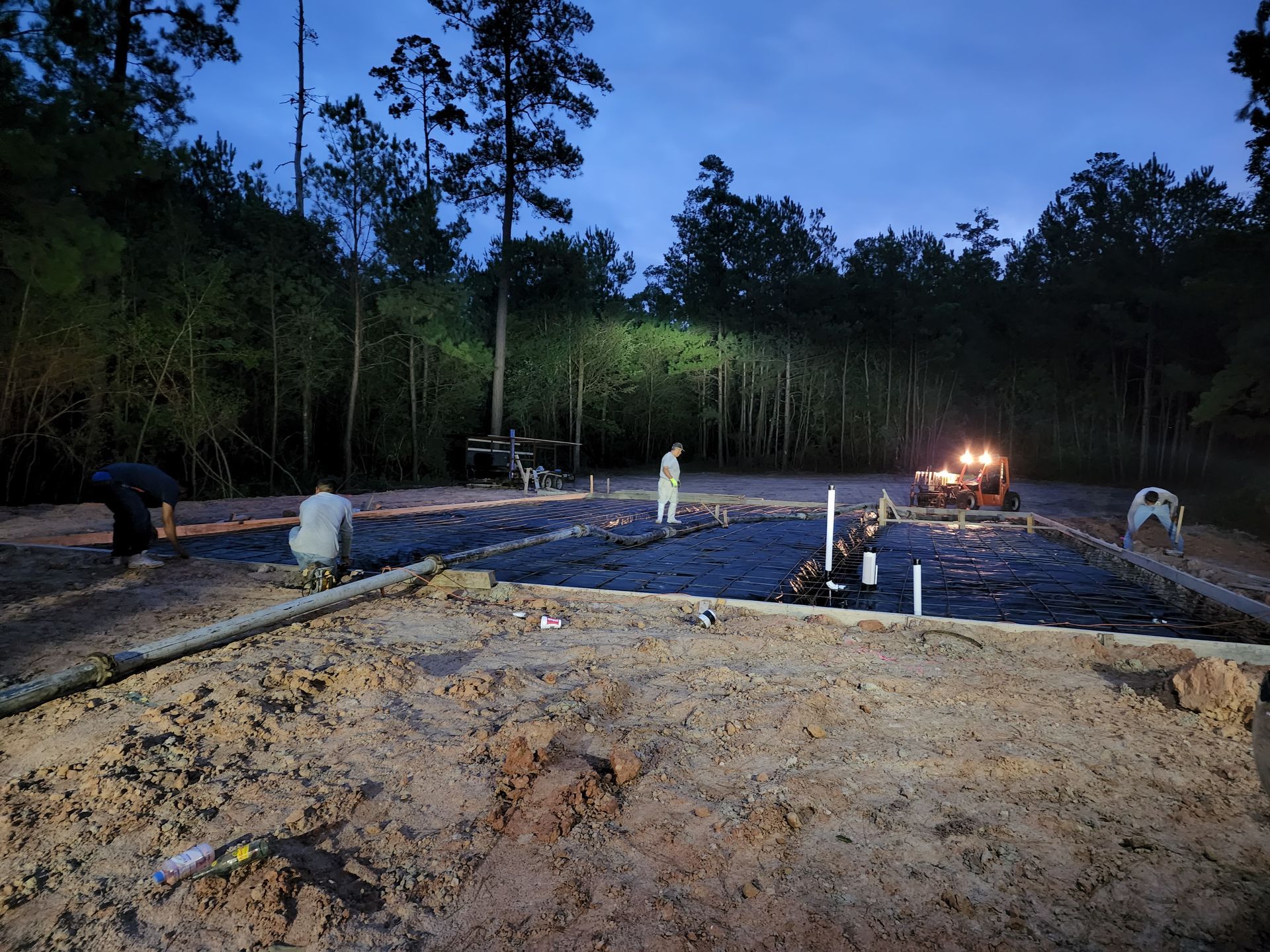 Construction site at dusk. Workers near a tractor, forming a foundation surrounded by trees.